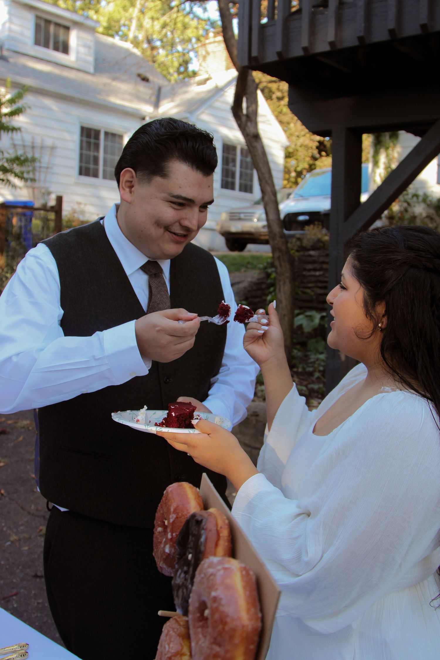 A couple shares bites of wedding cake with each other underneath a covered outdoor pavilion at sunset.