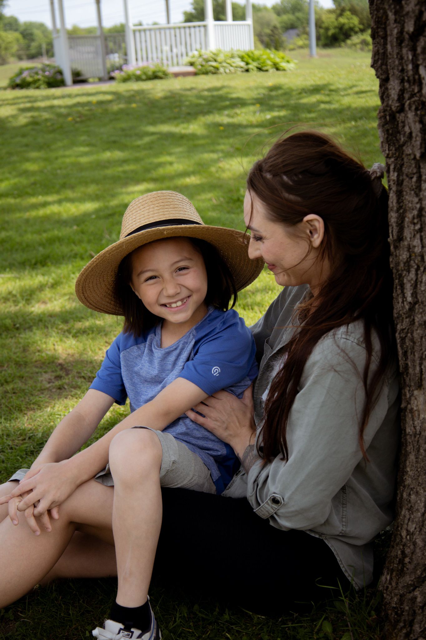 Two friends sit together under a shady tree on a sunny day in a grassy park area.