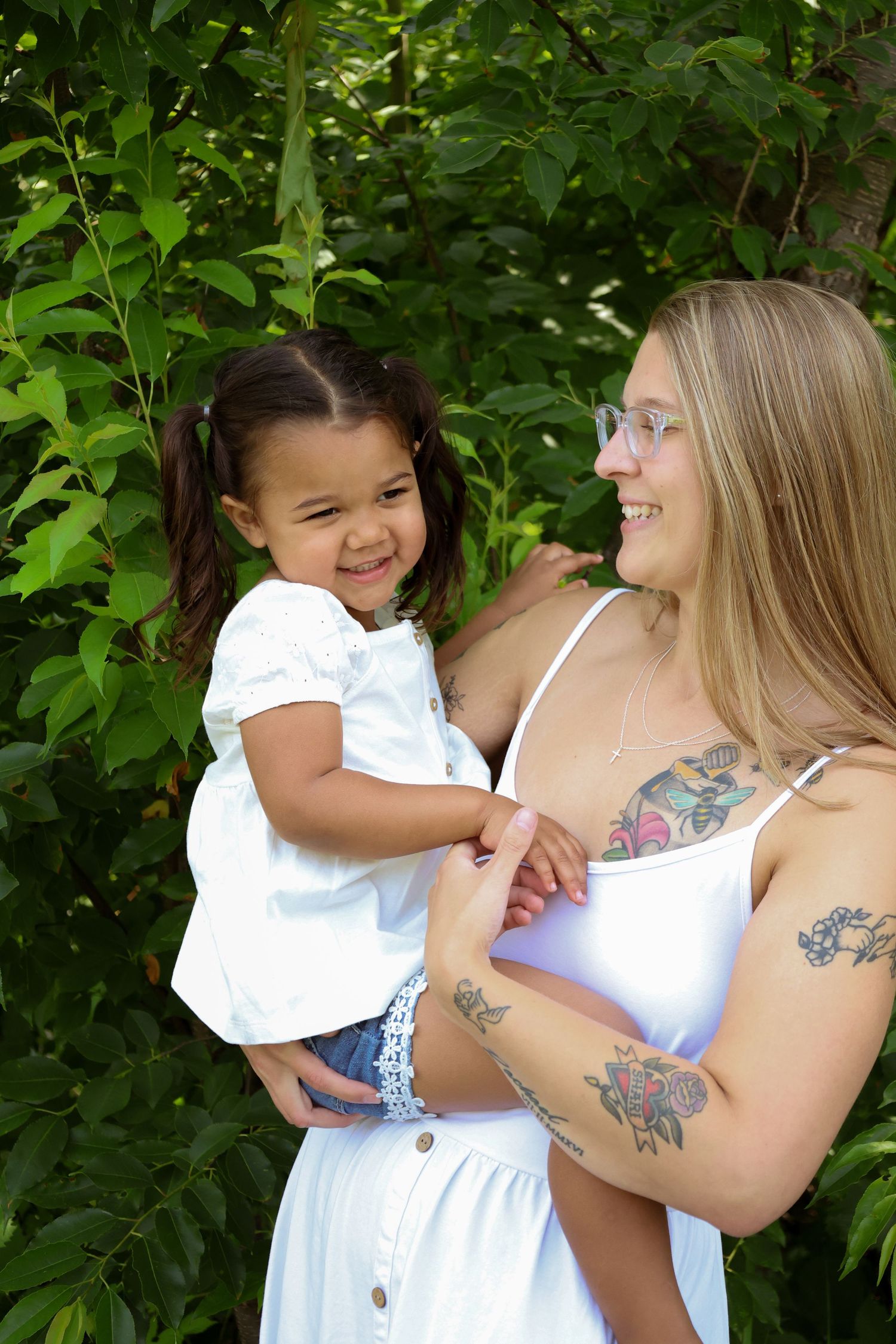A woman and toddler in white dresses share joyful laughter while embracing outdoors among green foliage.
