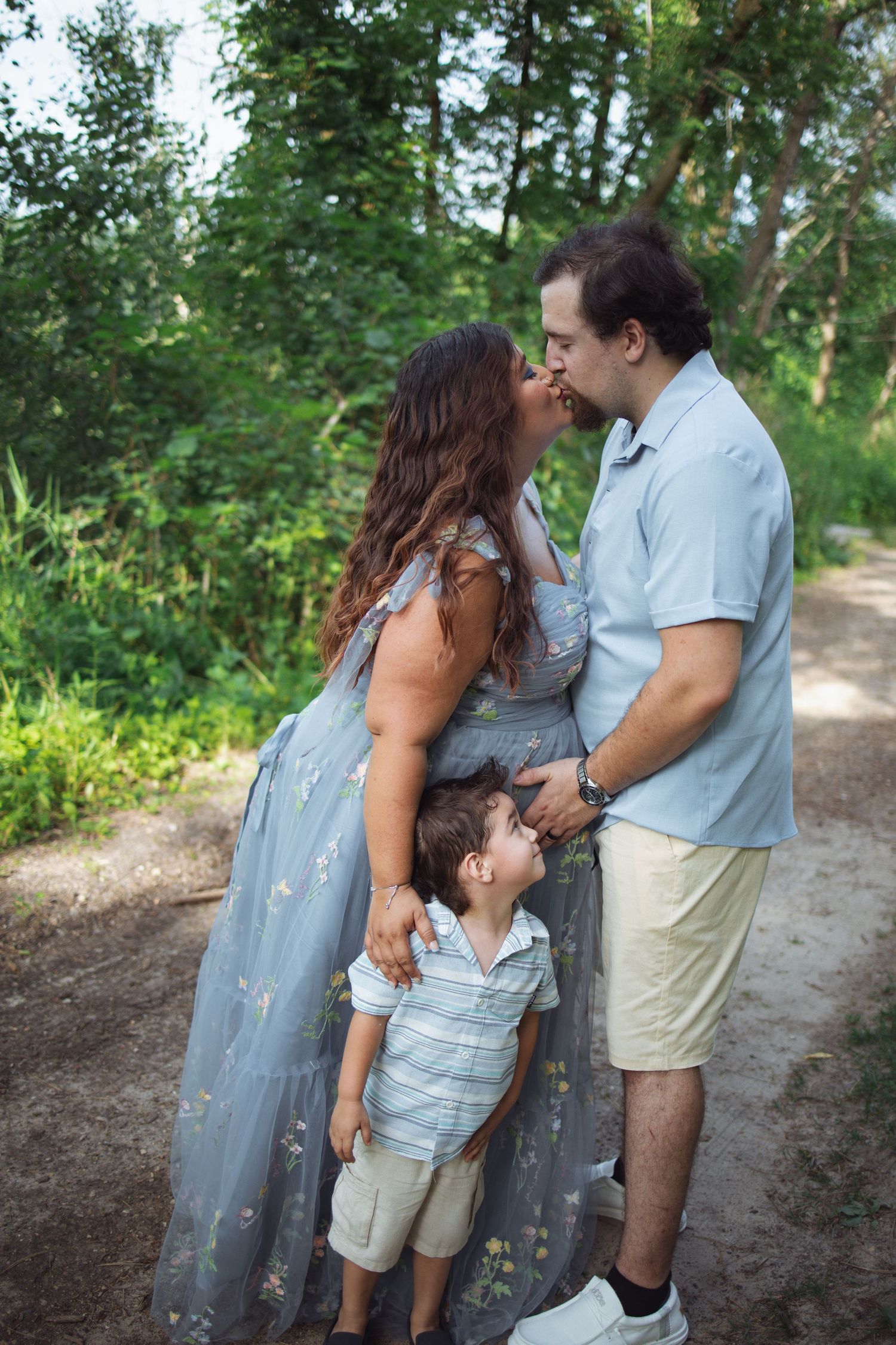 A couple and young child share tender moments together on a tree-lined dirt path on a sunny day.