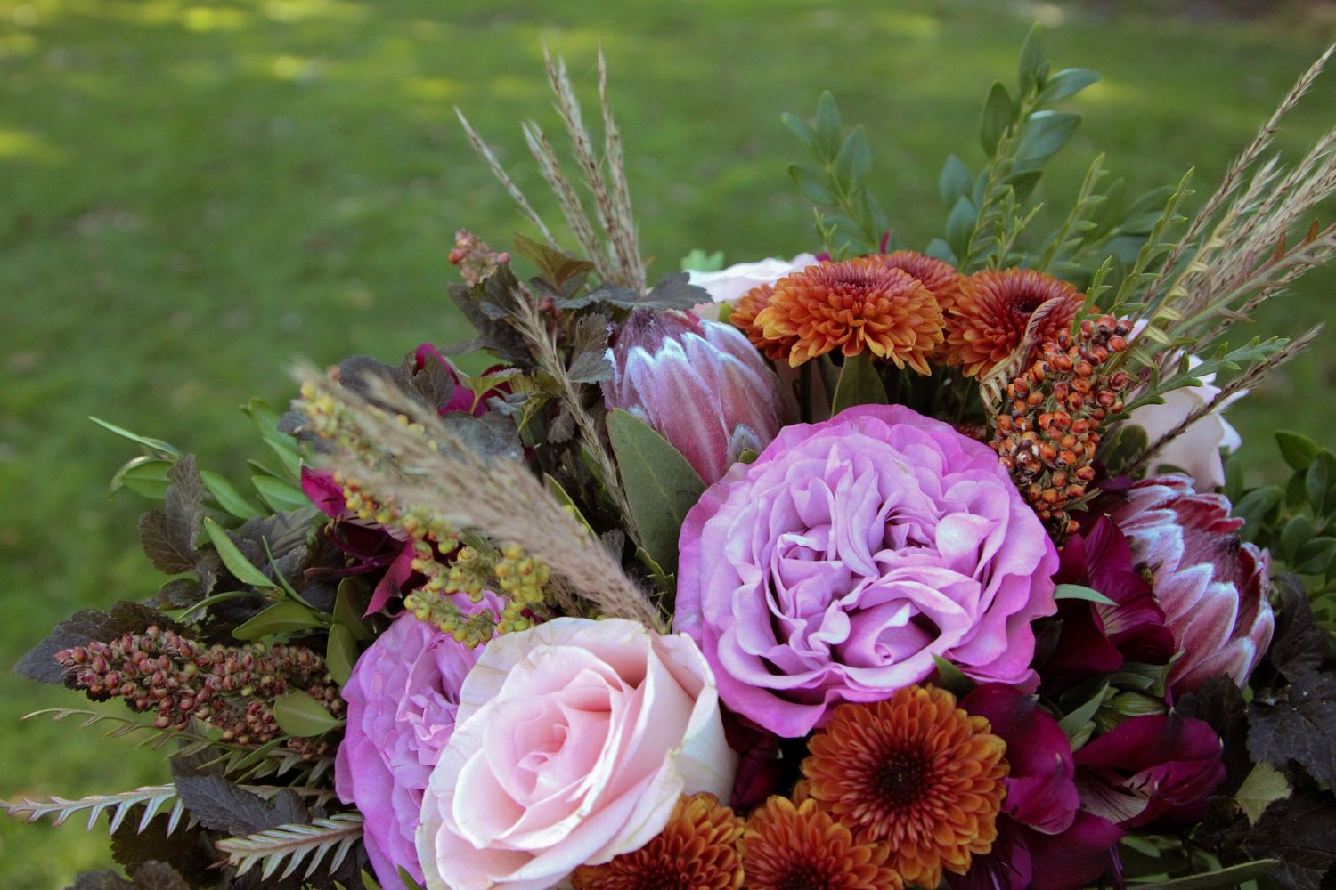 Beautiful autumn floral arrangement with pink roses, burgundy mums, and dried grasses against a blurred green background.
