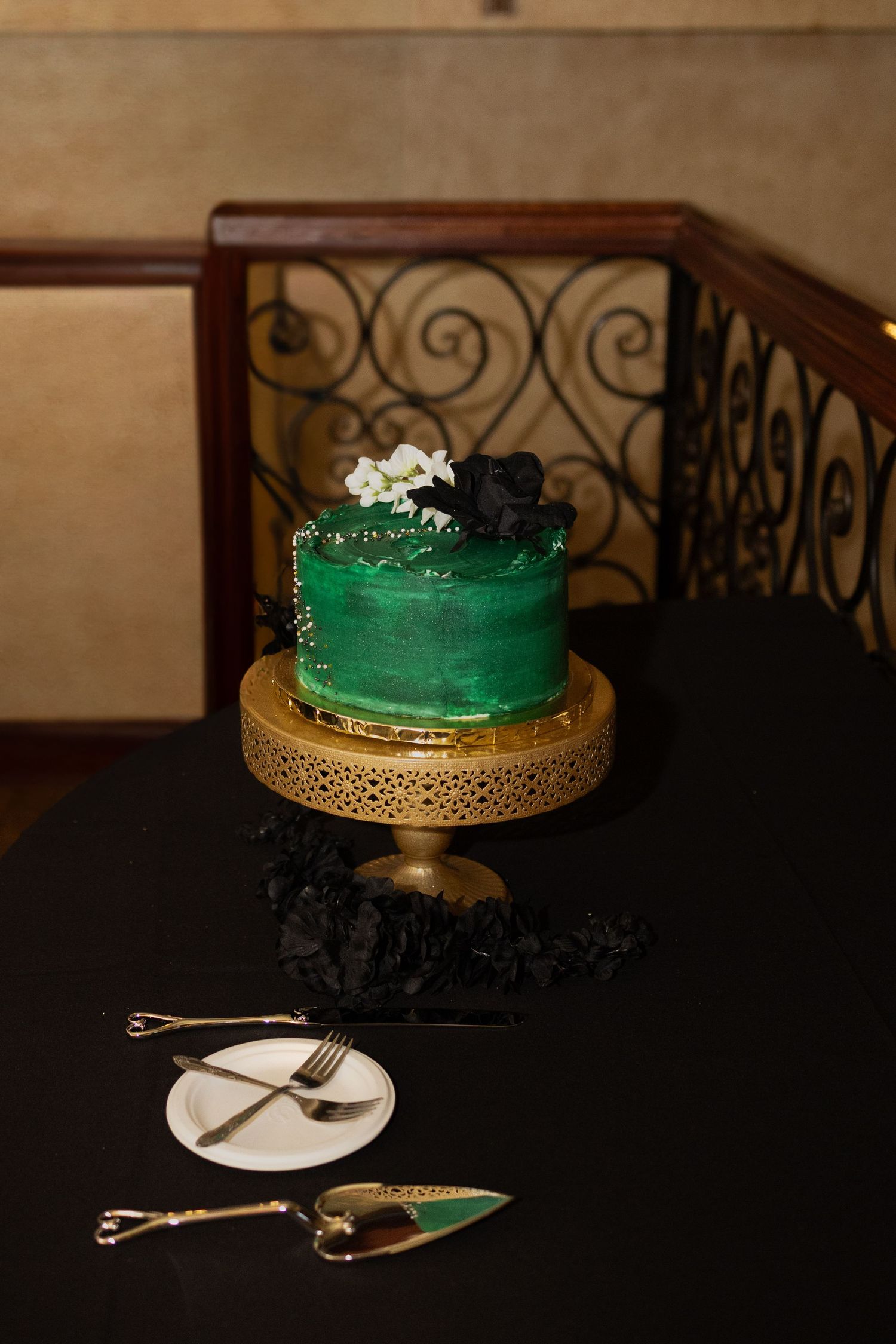 Elegant emerald green wedding cake with black accents on a gold decorative stand with vintage silverware on black tablecloth.