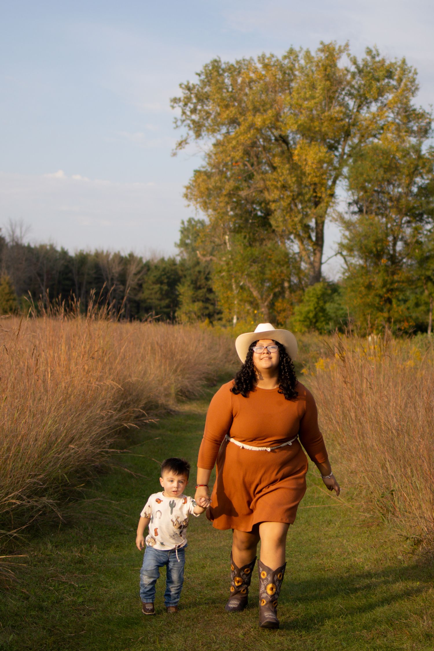 Two people walking together through a golden autumn field of tall grass at sunset with trees in the background.