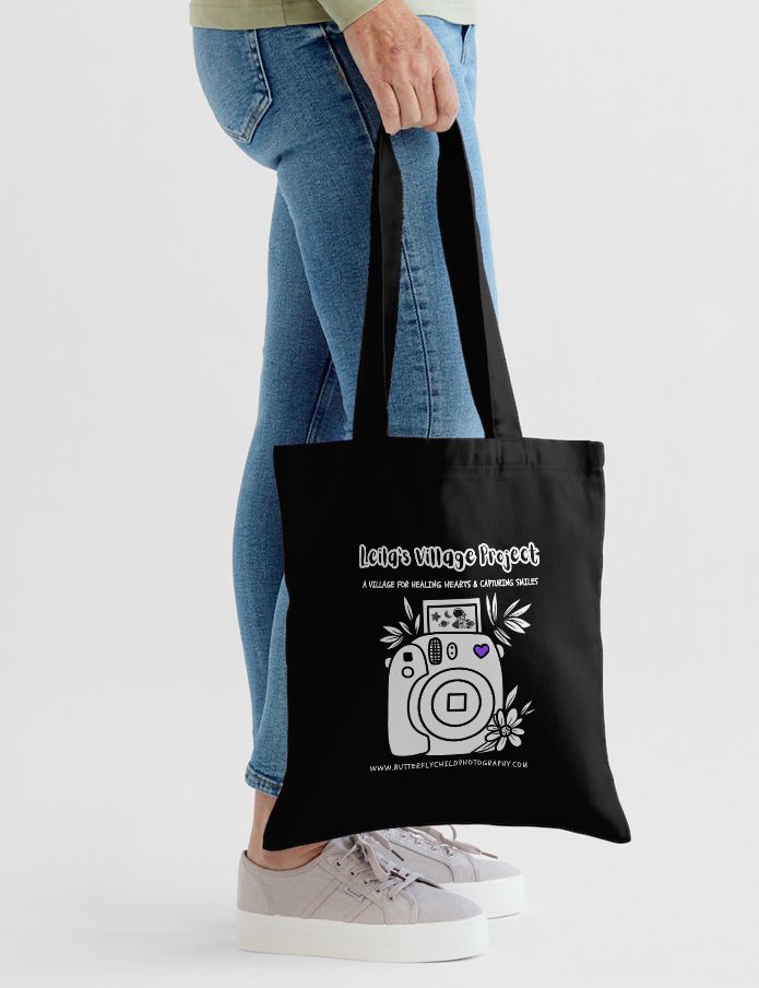 Black canvas tote bag with camera design and text being held against blue jeans and white sneakers.