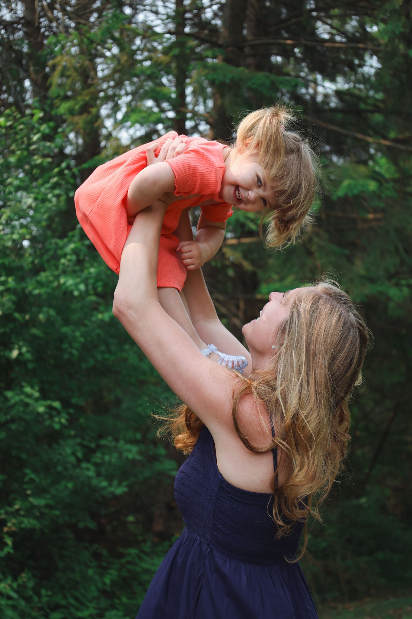 A touching moment outdoors as a parent lifts a young child wearing coral dress against a green forest background.