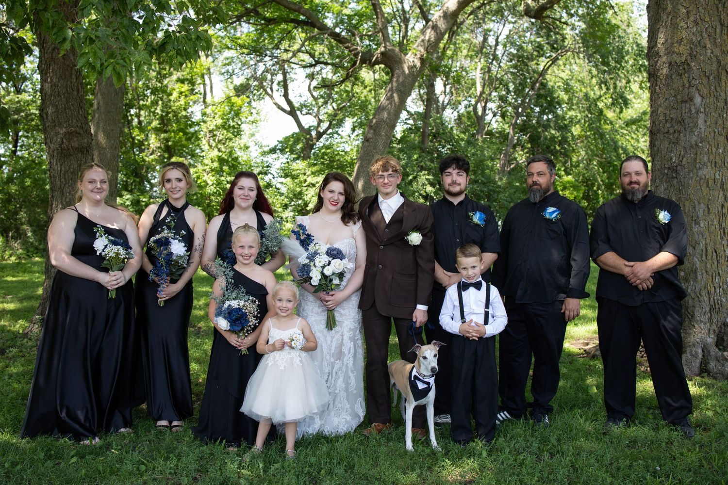 Wedding party poses outdoors in formal attire with black dresses, suits and a spotted dog under green trees.
