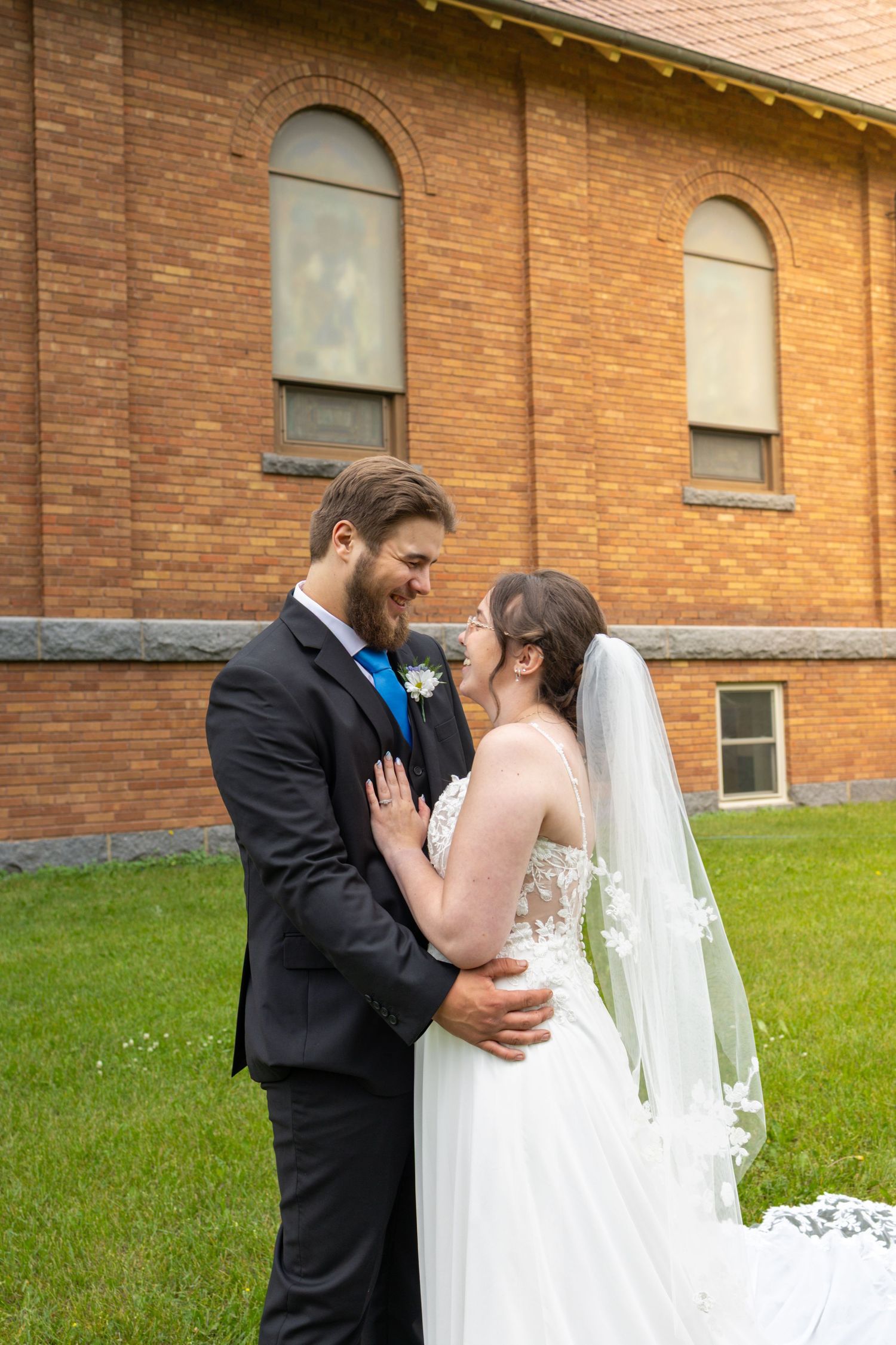 A couple in wedding attire embrace outside a brick church building with arched windows.