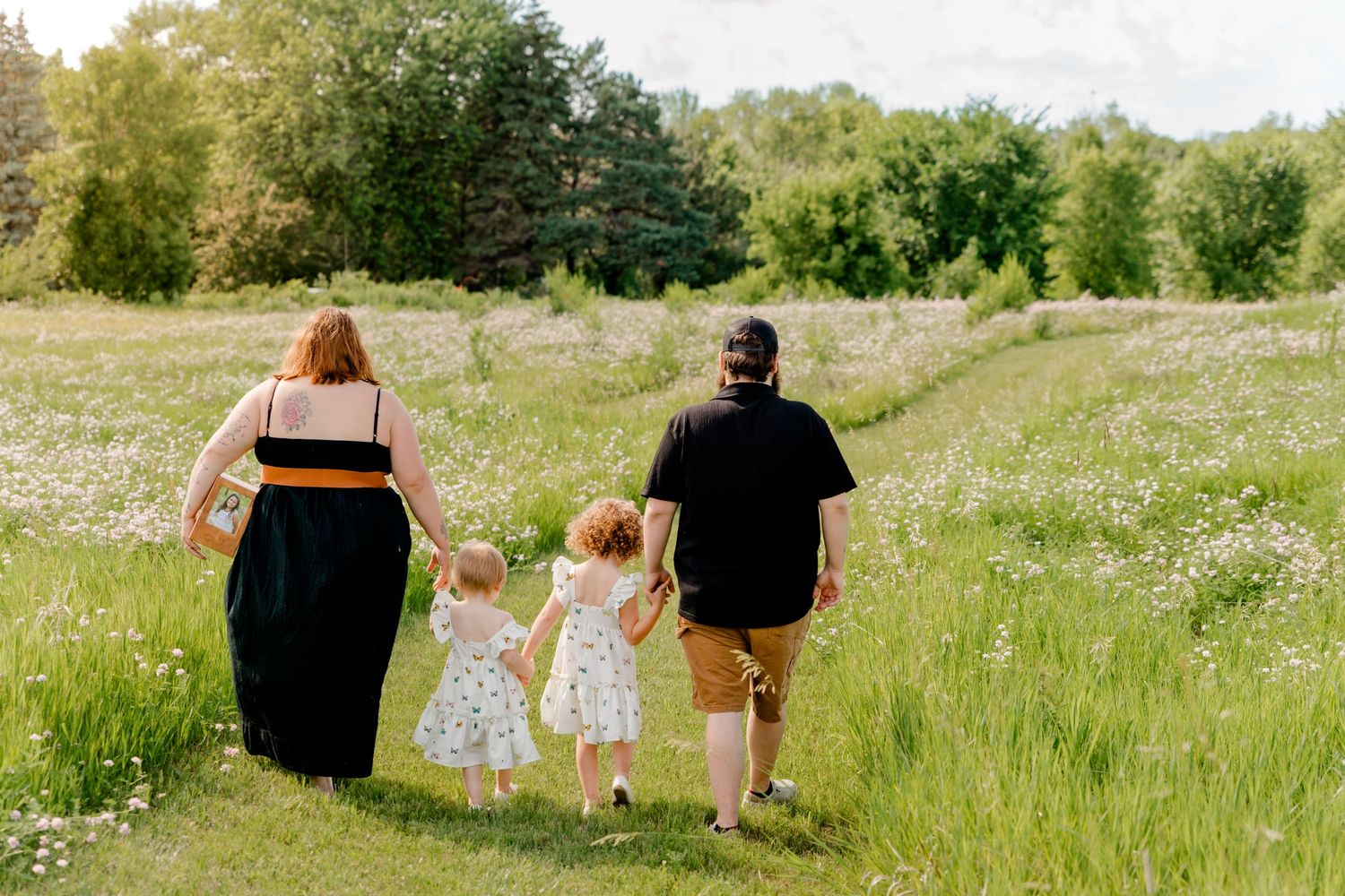 Family walking hand in hand through a sunlit meadow with wildflowers and trees along a grassy path on a summer day.