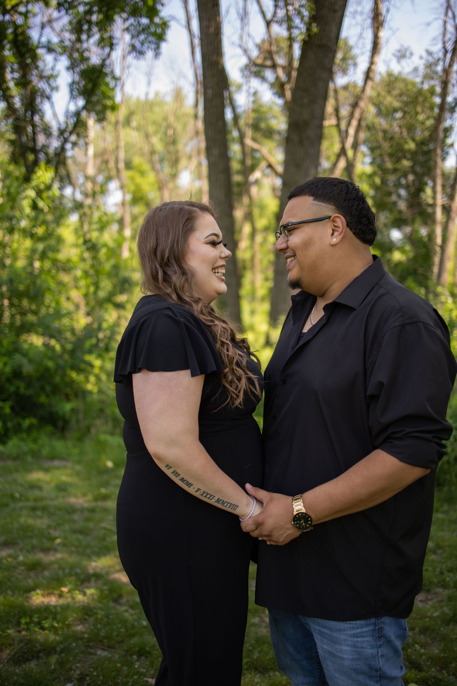 A couple in black attire share intimate moments of laughter and embrace in a sunlit wooded setting.