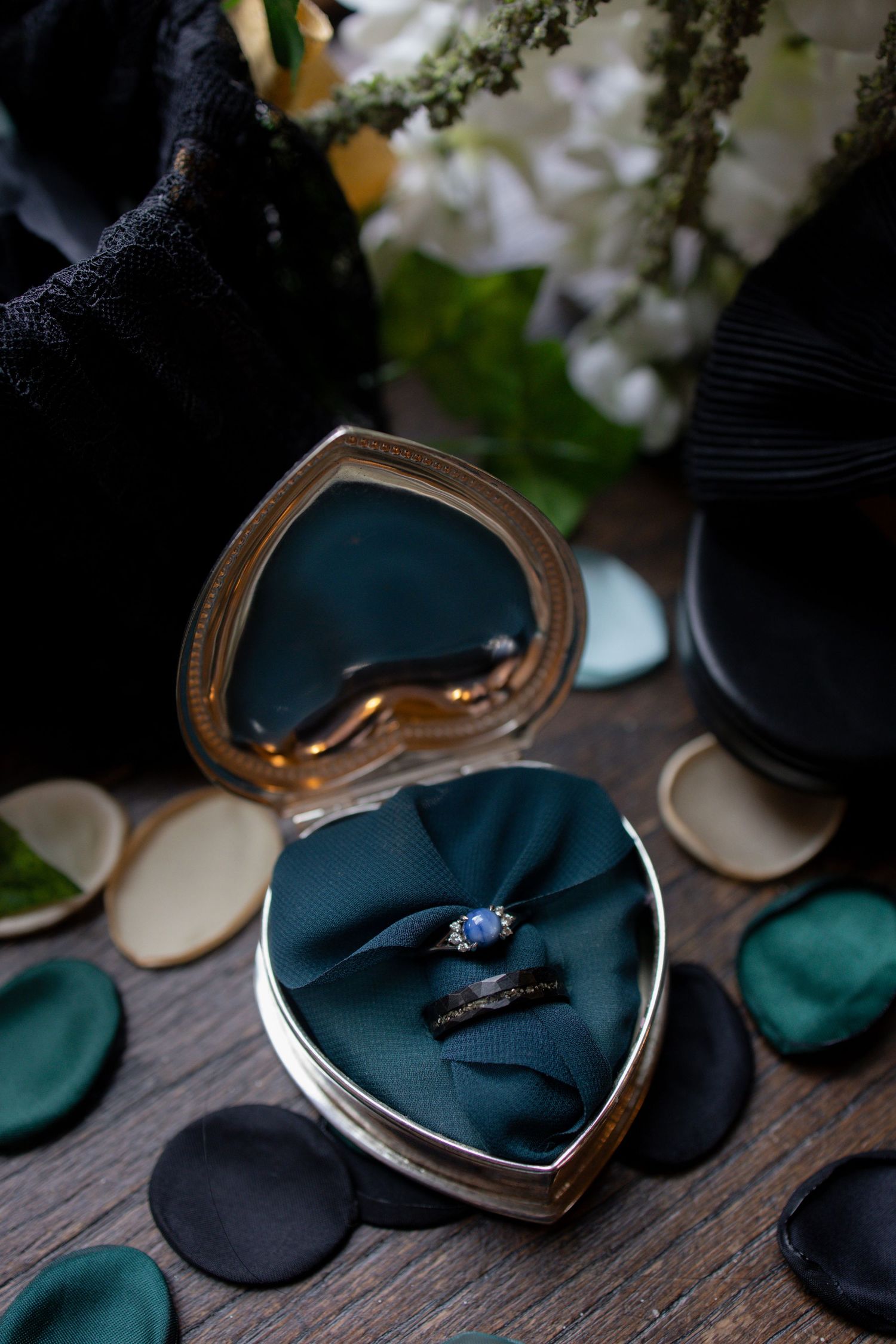 Wedding rings displayed in a heart-shaped emerald velvet jewelry box with scattered dark fabric circles on wooden surface.