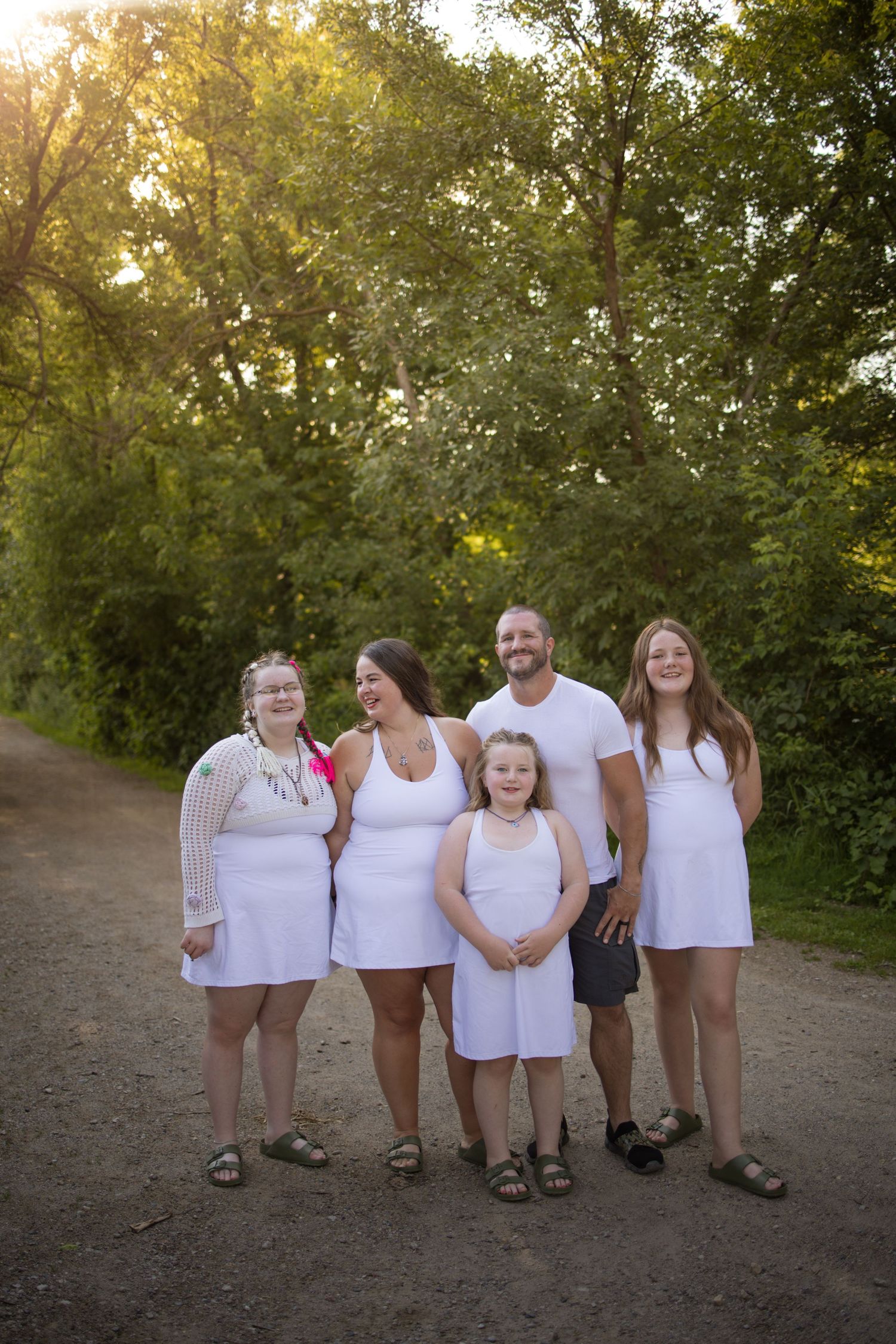 A group wearing matching white tops poses on a dirt path with green trees in the background at sunset.