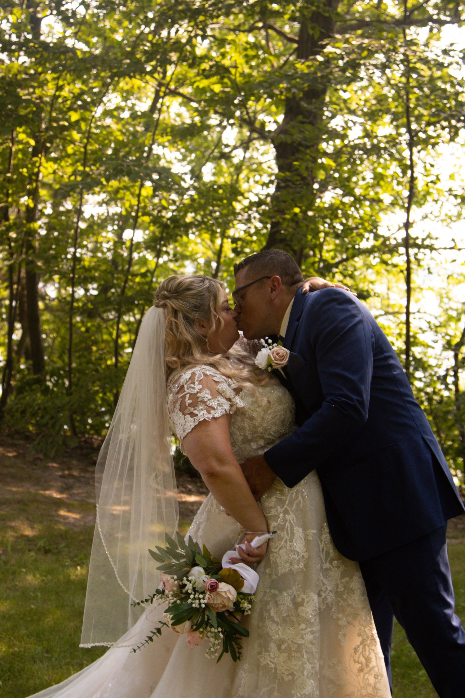 Newlyweds share a romantic kiss in a sunlit forest setting while holding a floral bouquet.
