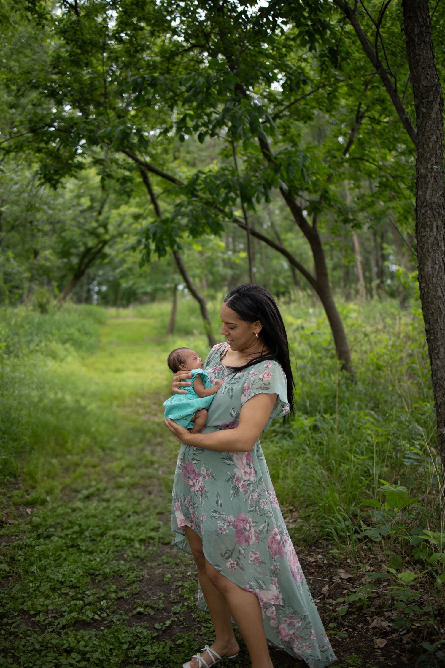A person in a floral dress holds a baby while standing on a path in a lush green woodland setting.