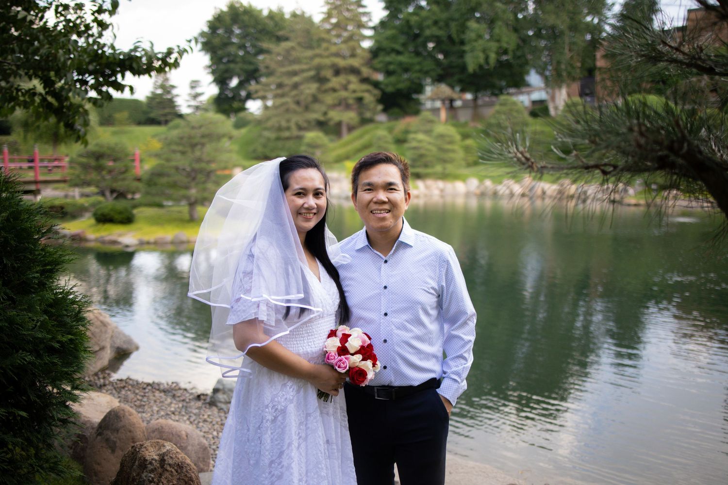 A romantic wedding couple poses together by a serene lake, with trees and greenery in the background.