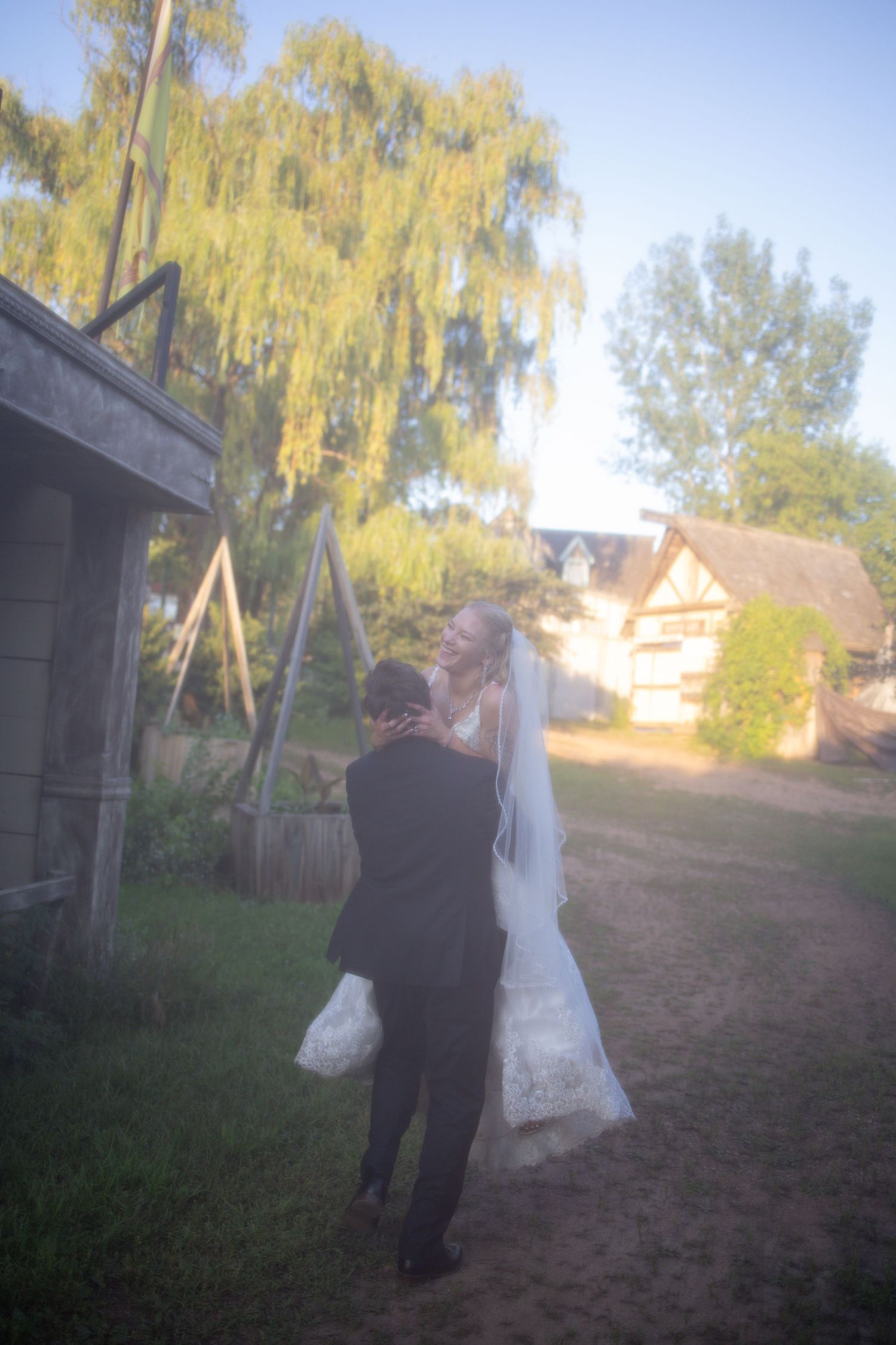 A couple walks along a dirt path under a weeping willow tree at a rustic outdoor wedding venue.