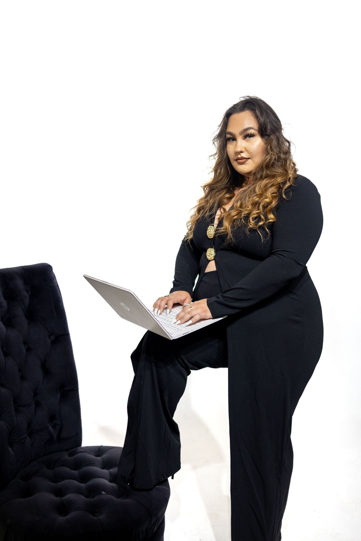 Professional woman in black business attire works on laptop while standing next to dark tufted chair against white backdrop.