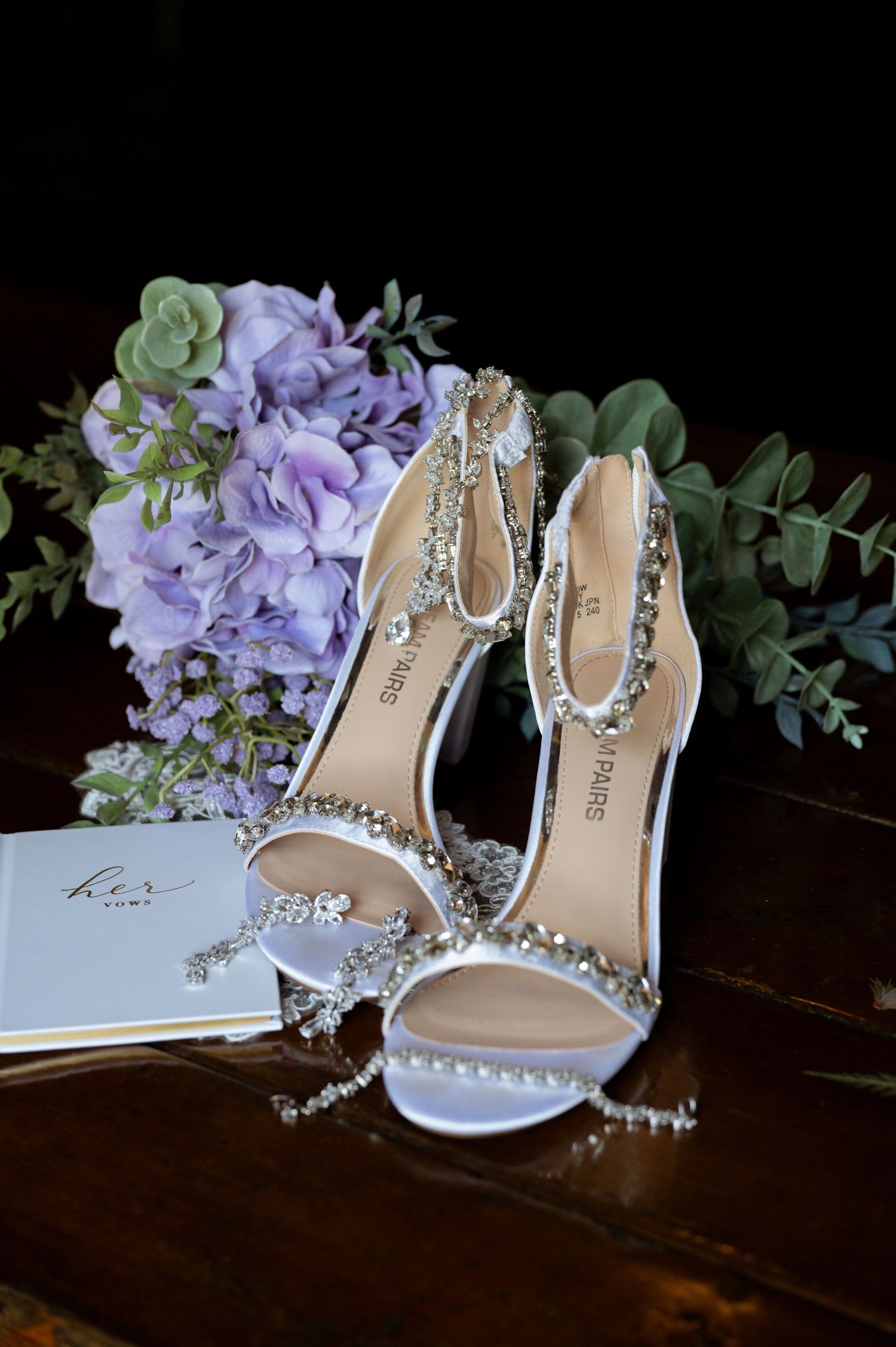 Crystal-embellished white bridal heels displayed with delicate purple hydrangeas and greenery on dark wooden surface.