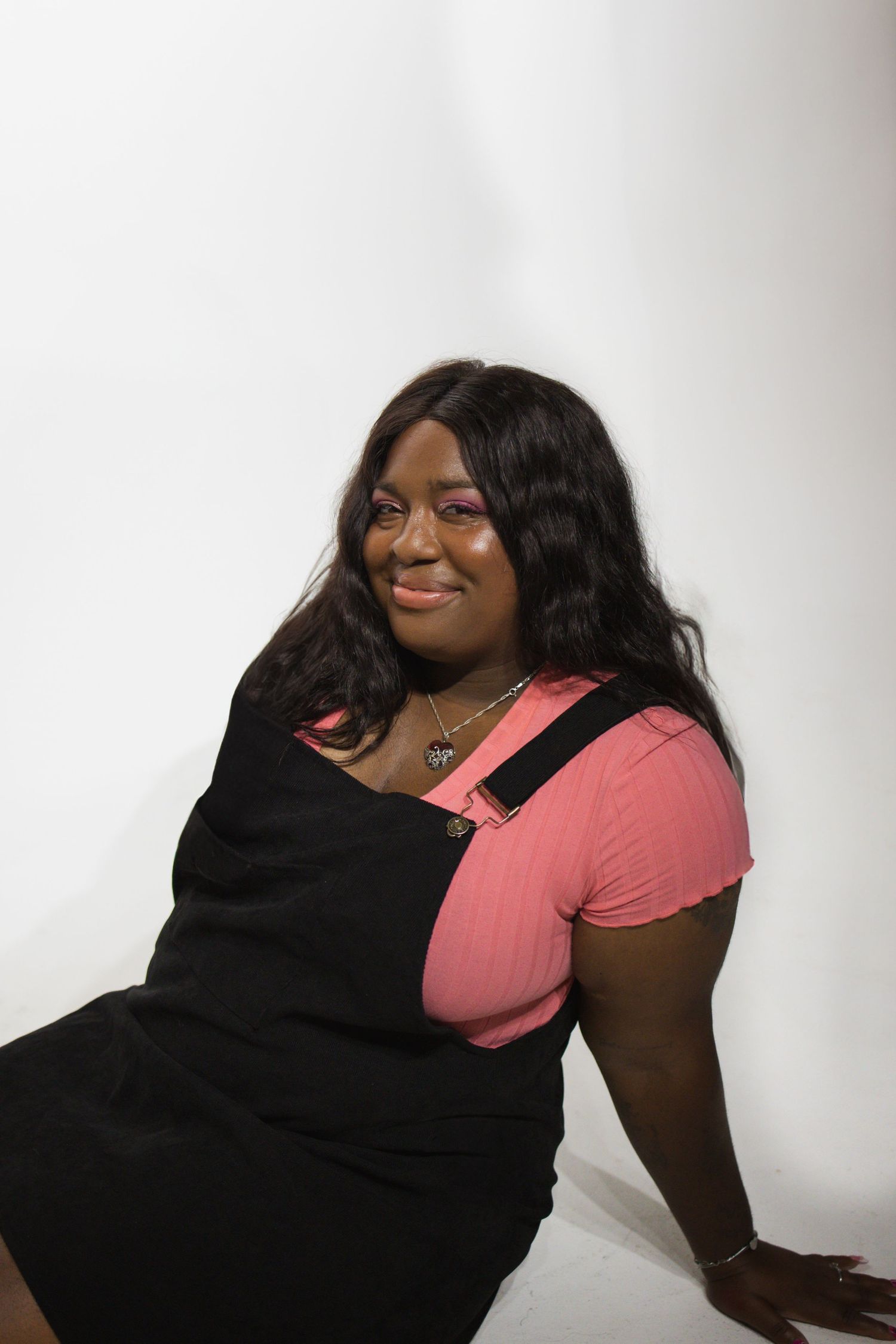 Person wearing a pink t-shirt and black overall dress poses against a white backdrop for a portrait.