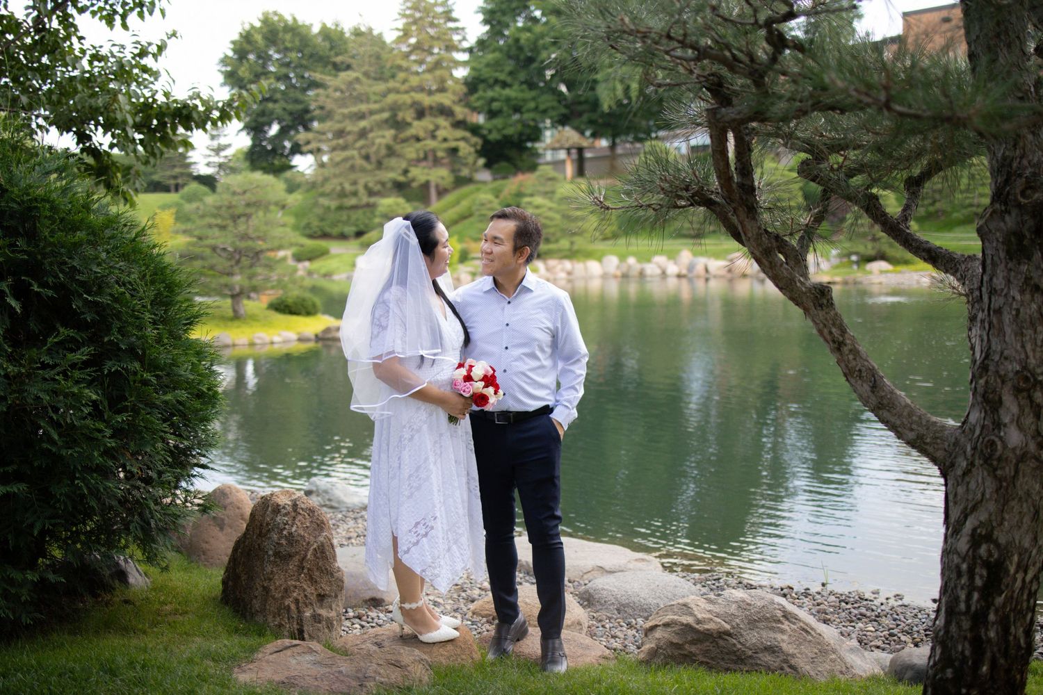 A couple in white robes shares a romantic moment by a serene lake surrounded by lush greenery.