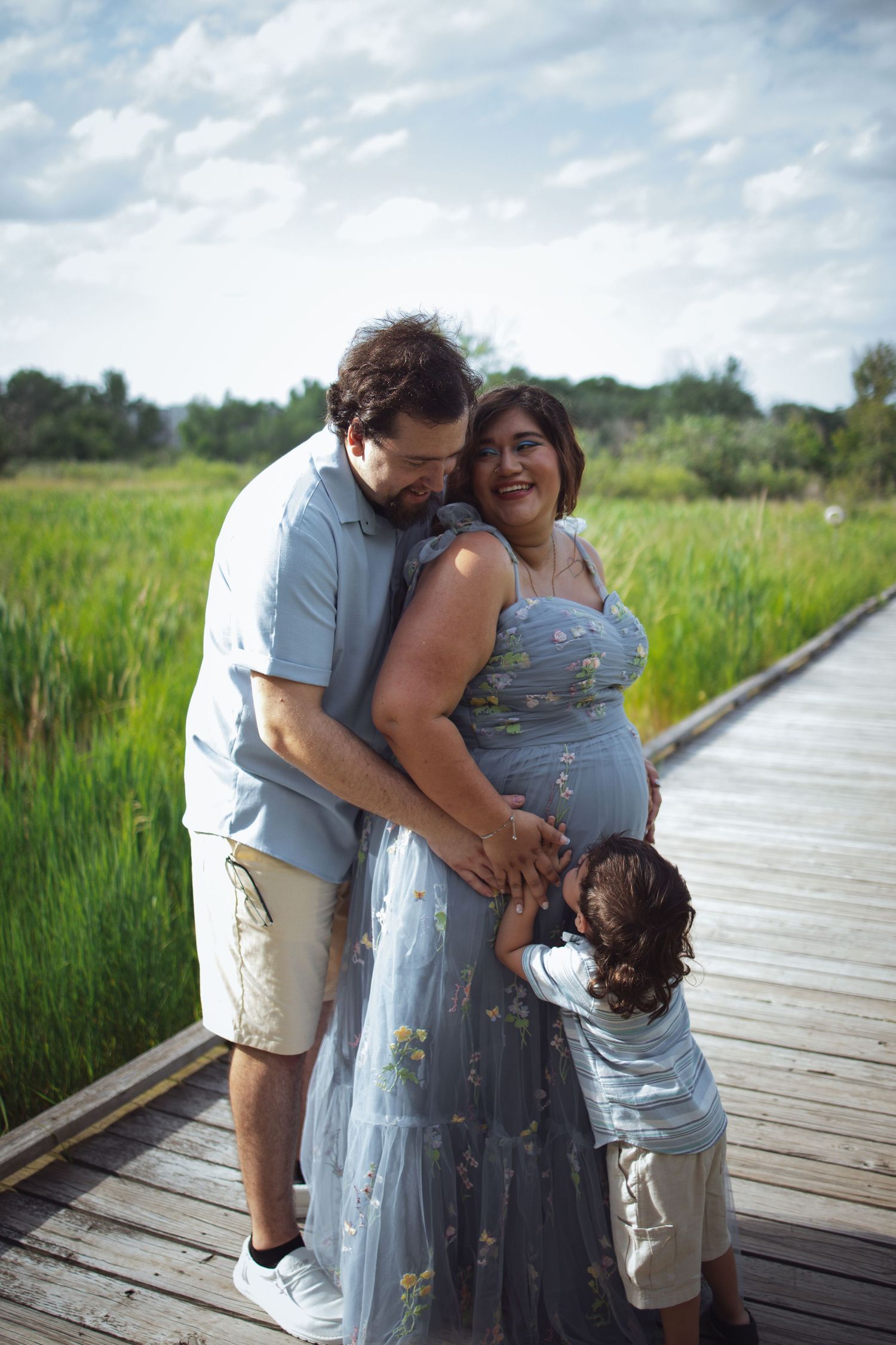 Family shares a tender moment together while standing on a wooden boardwalk surrounded by tall marsh grasses on a cloudy day.