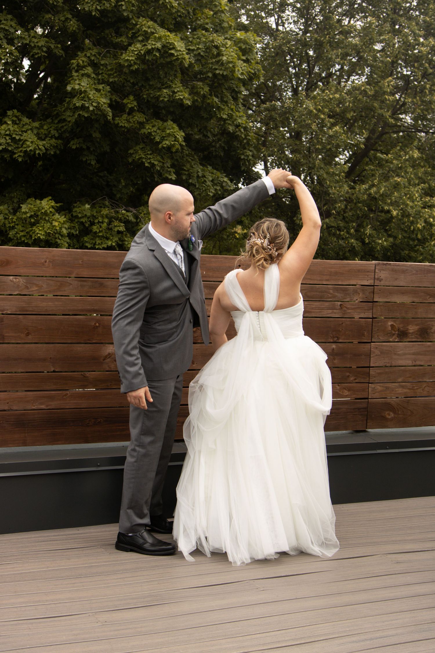 A couple in wedding attire dances together on a wooden deck surrounded by trees in the background.