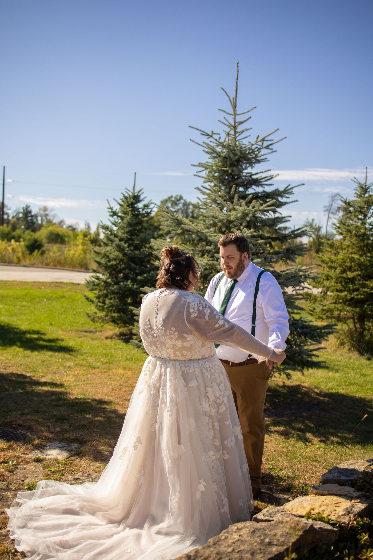 A couple in wedding attire share a tender moment among evergreen trees on a sunny autumn day.