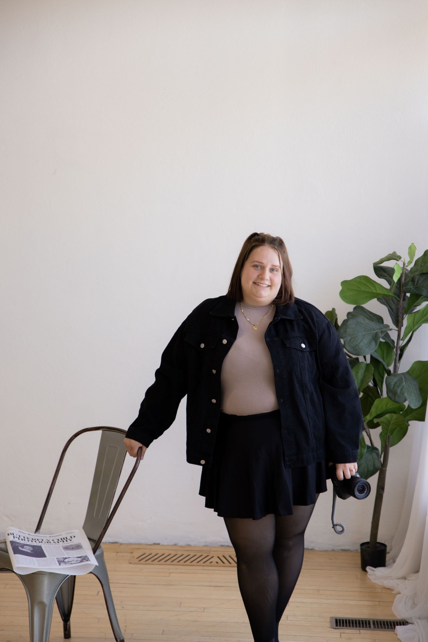 A person in a black denim jacket and beige top poses with a chair in a minimal room with a fiddle leaf fig plant.