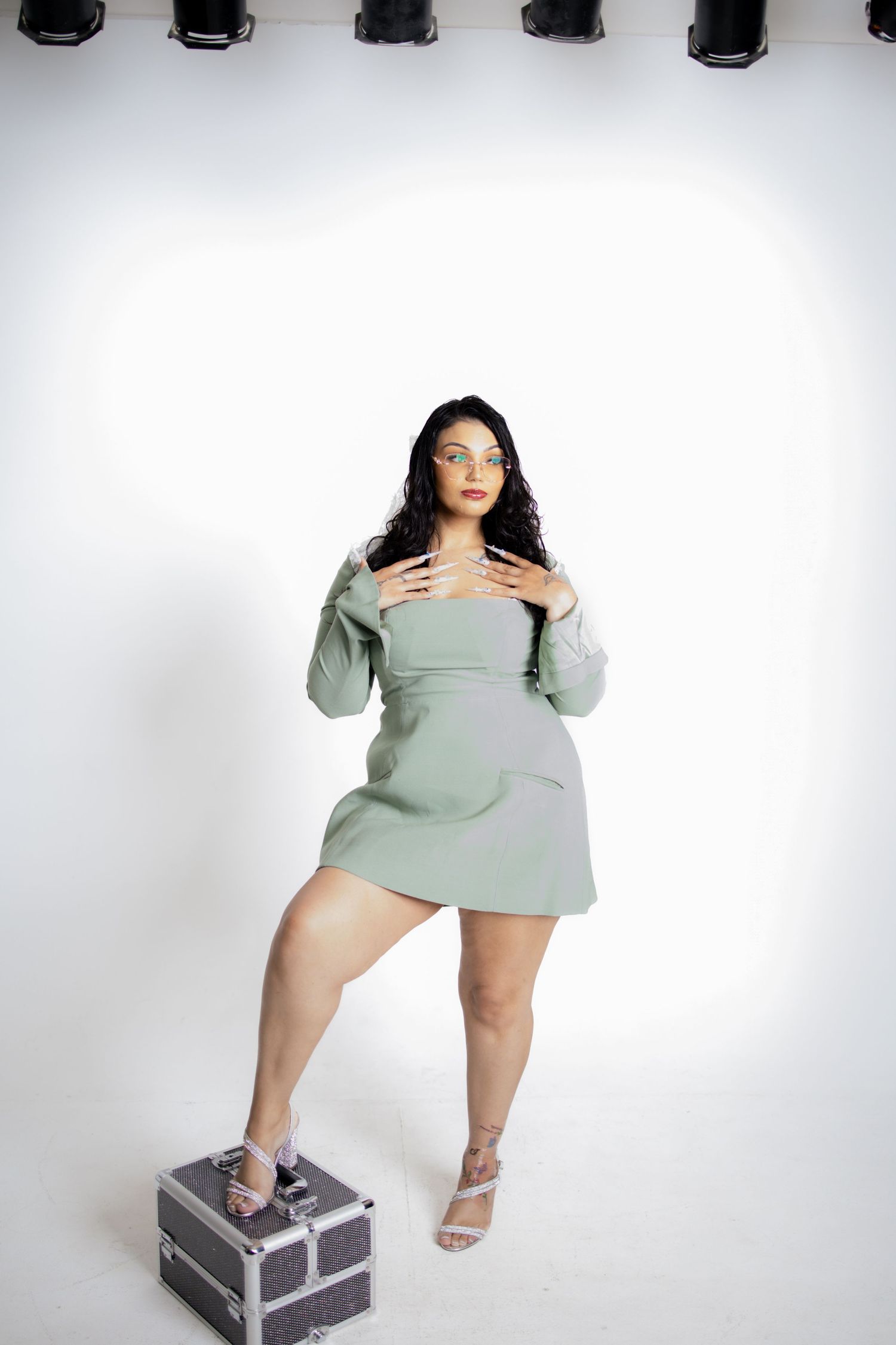 Figure in pale mint green sweater dress poses dynamically on gray equipment case against white background.