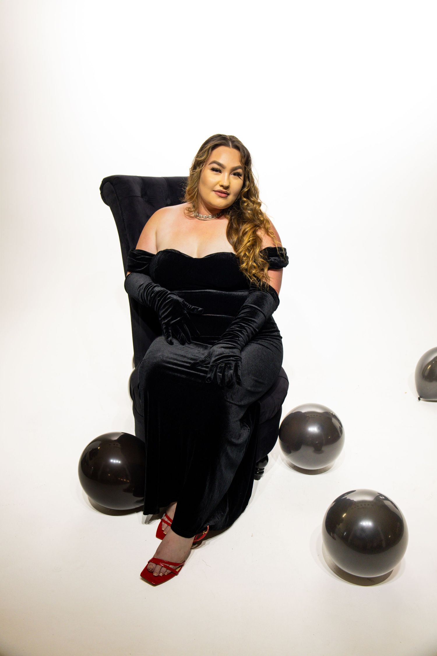 Model in black velvet dress poses artistically with dark spherical props against white backdrop.