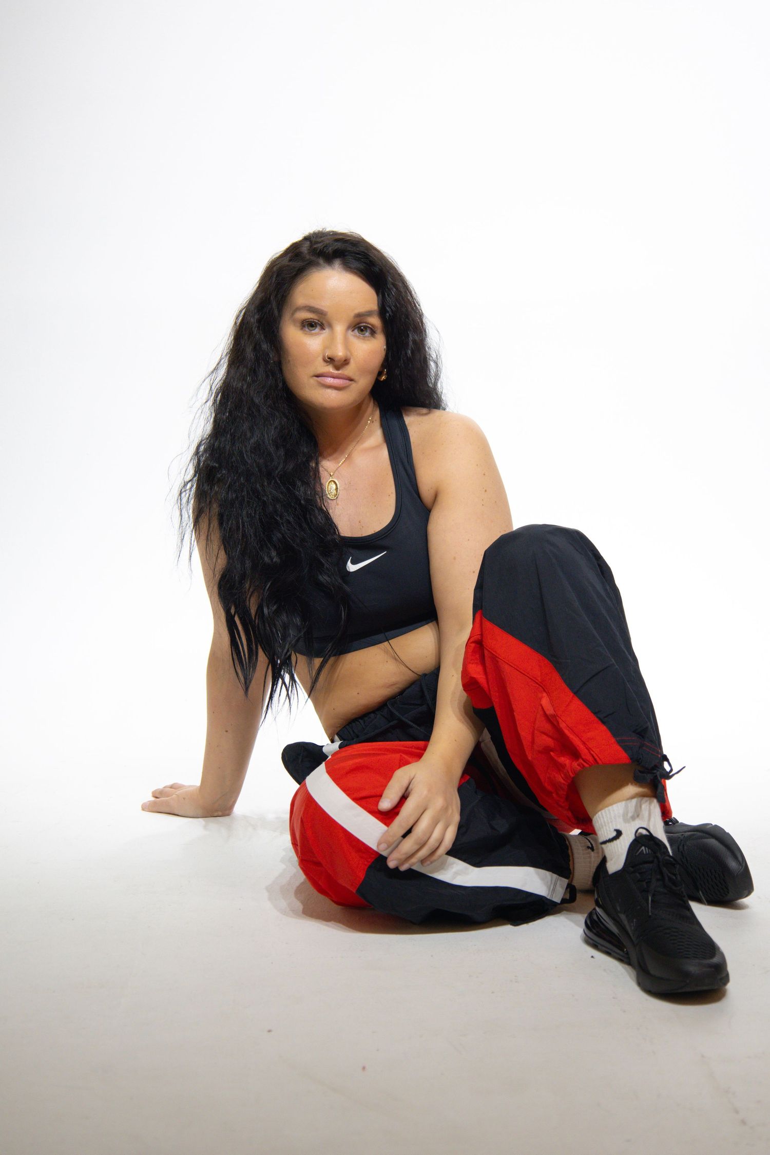 Athletic woman in Nike sports bra and red-black track pants sits on white floor in casual pose.