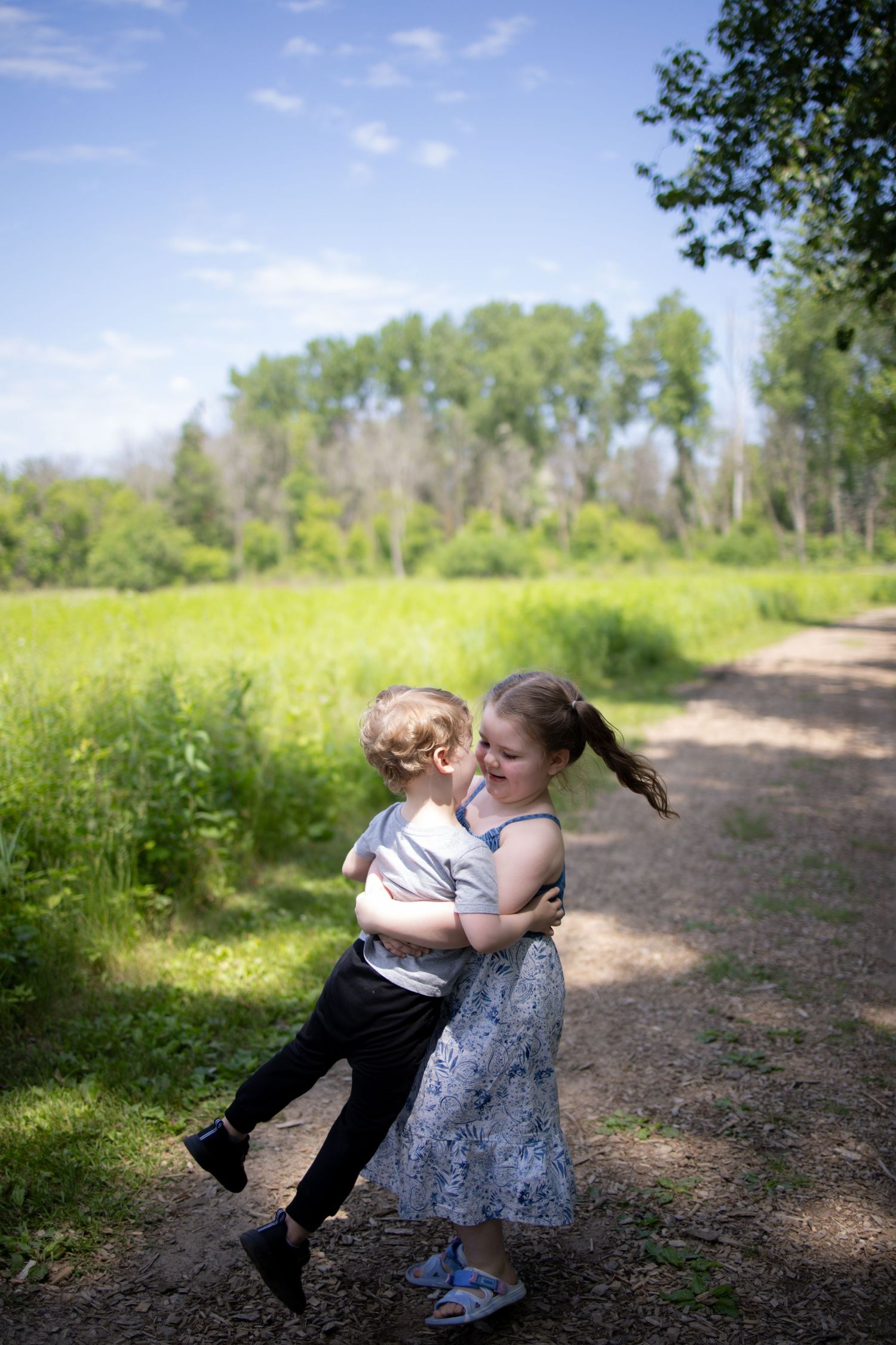 Two young children sharing an embrace while standing on a dirt path surrounded by lush green grass and trees in summer.