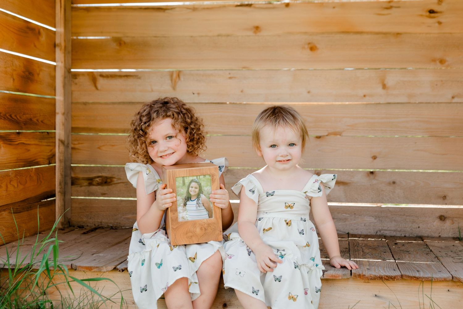 Two small children in floral dresses sit on a wooden porch holding a picture book against rustic wooden walls.