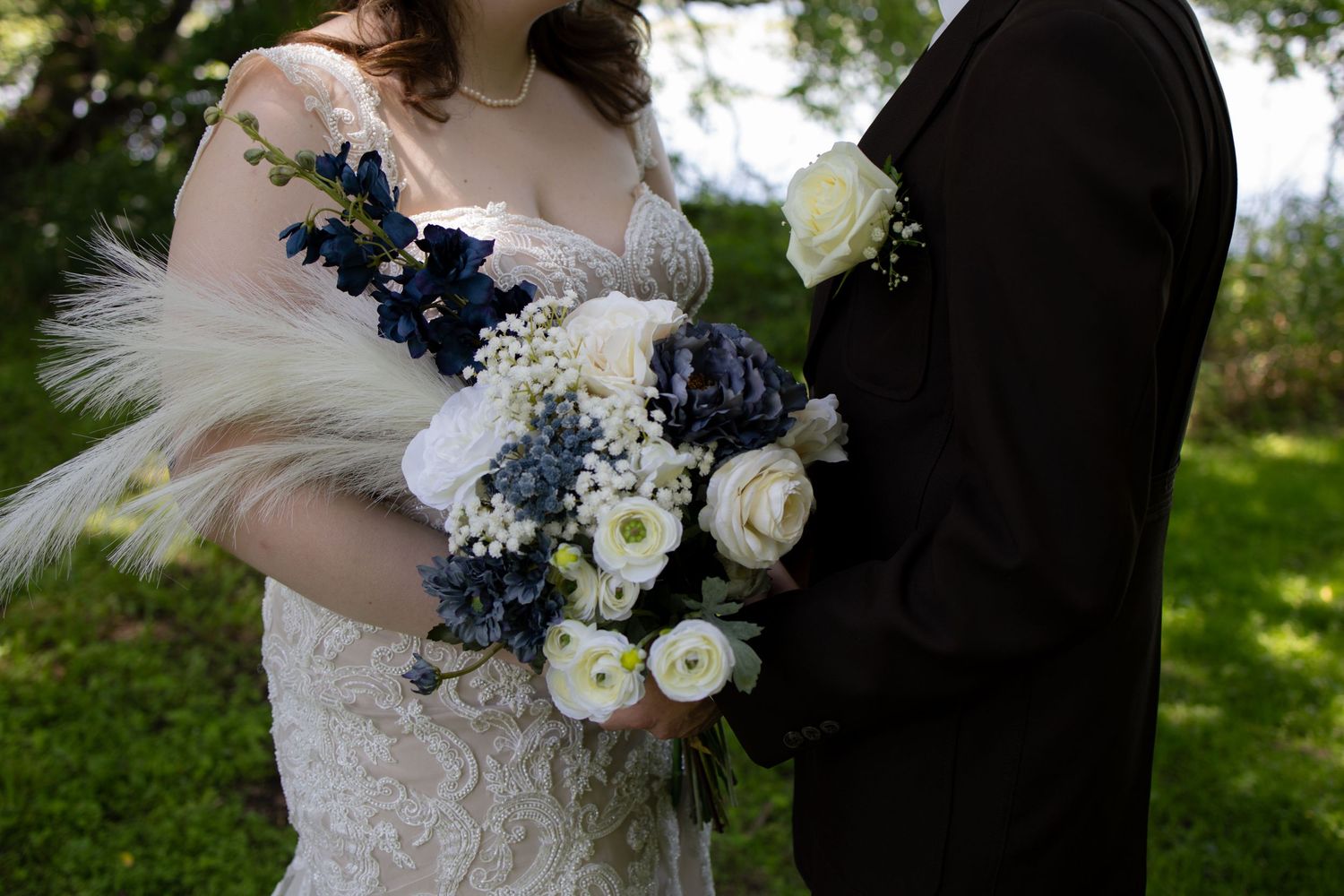 A close-up detail of white wedding dress lace and navy blue floral bouquet accents against a natural outdoor background.