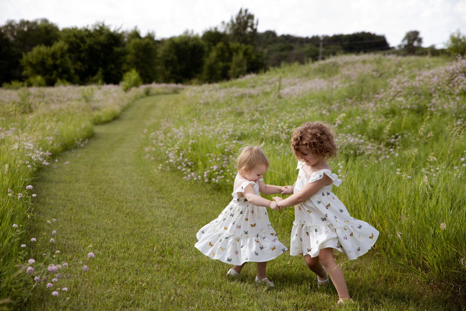 Two figures in flowy white dresses dance playfully together in a sunny summer meadow filled with tall grass.