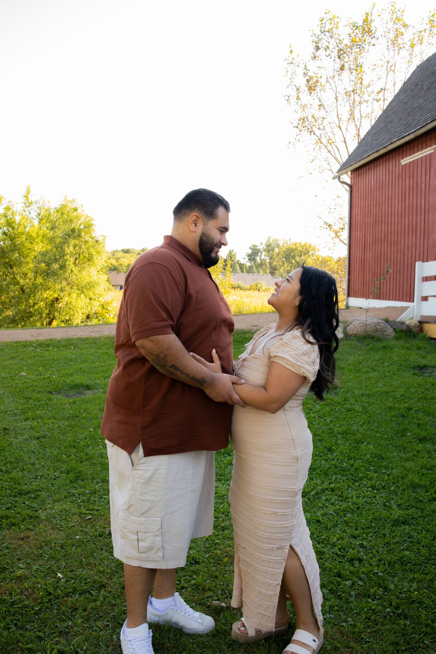 A couple shares a romantic moment together in front of a red barn on a grassy field during golden hour.