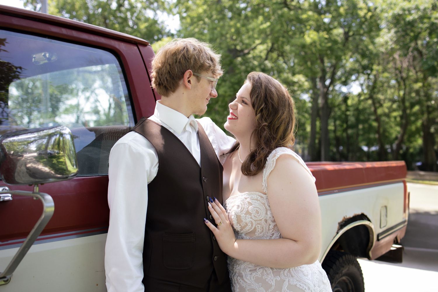A happy couple shares a romantic moment in front of a vintage red pickup truck in a sunlit outdoor setting.