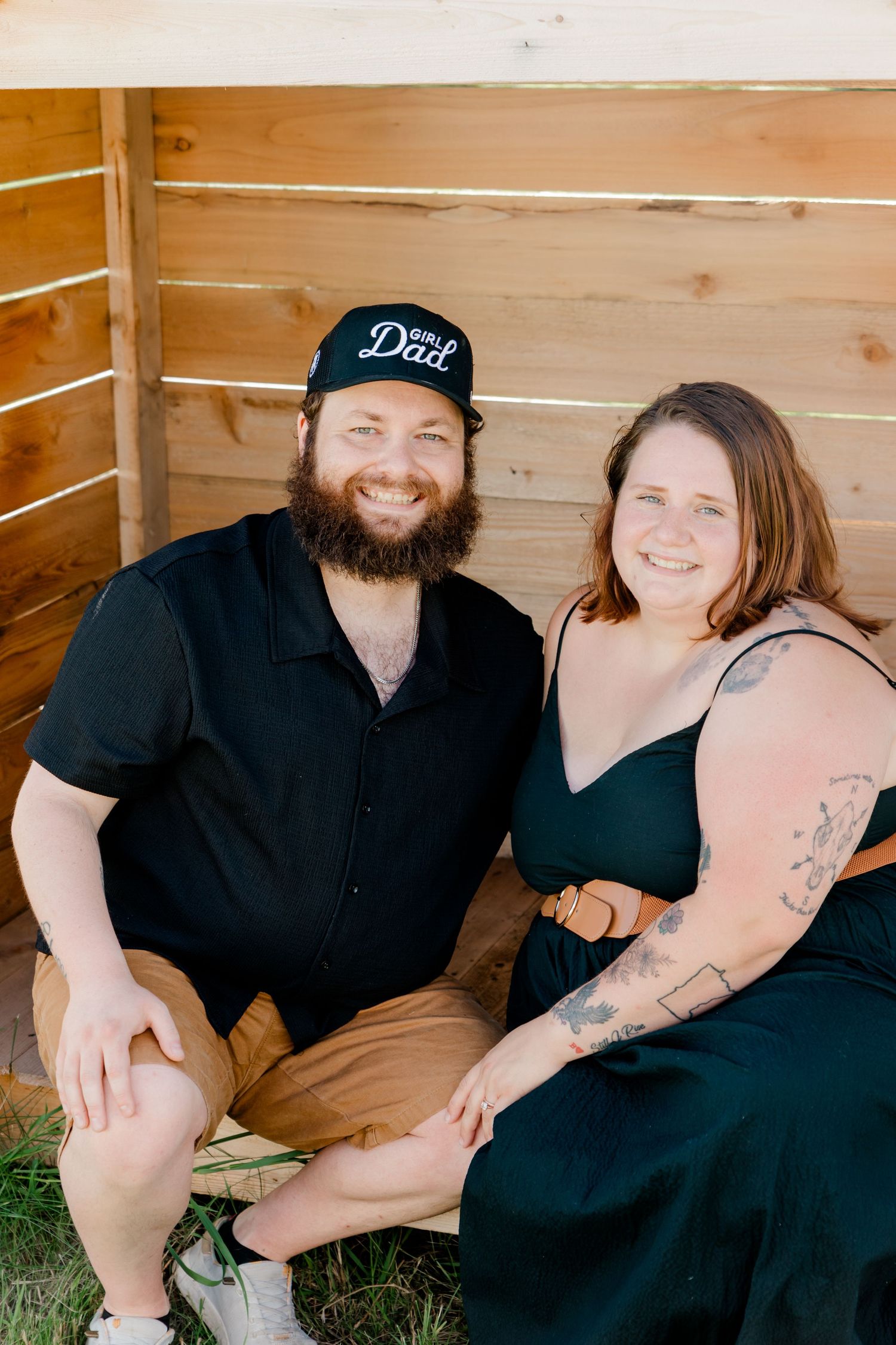 Couple in casual black attire sitting together against wooden wall background.