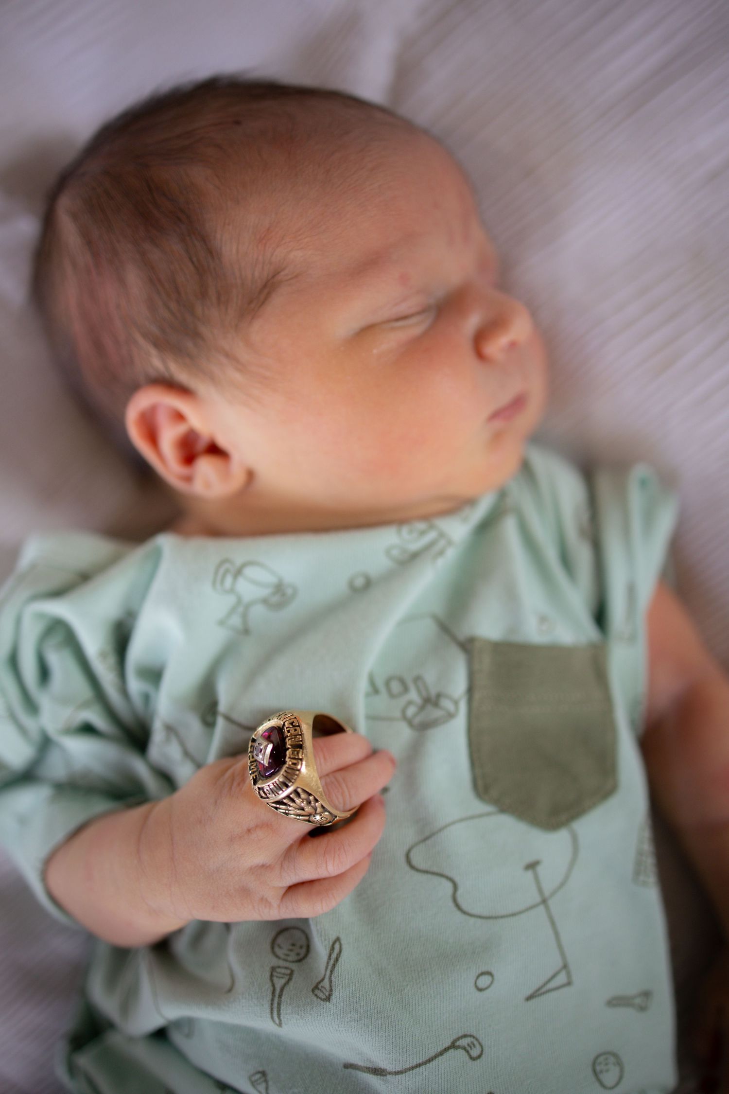 A sleeping baby in mint green clothing wears a large decorative ring on their tiny hand.