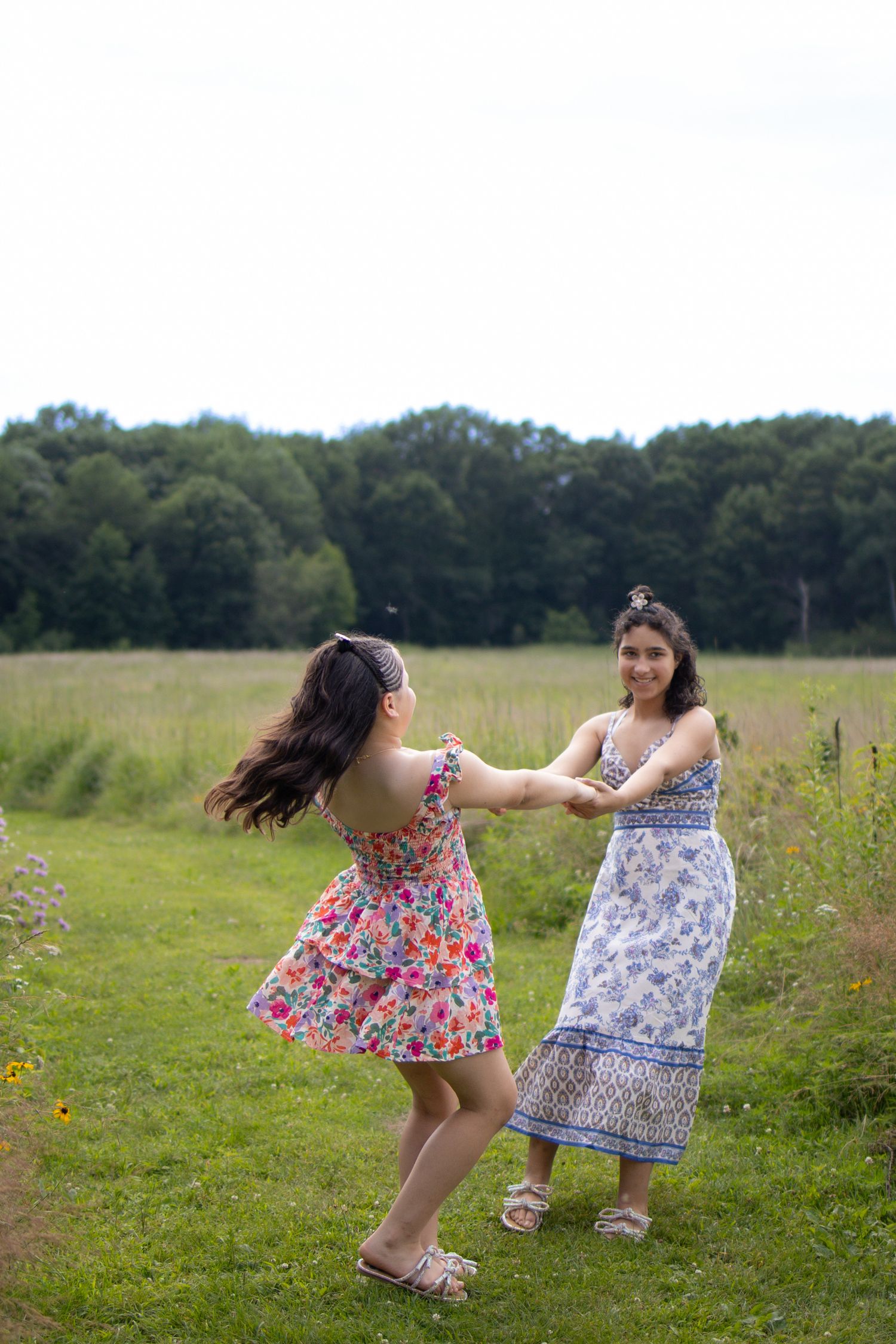 Two friends in summer dresses dance together in a sunny meadow surrounded by green trees and wildflowers.