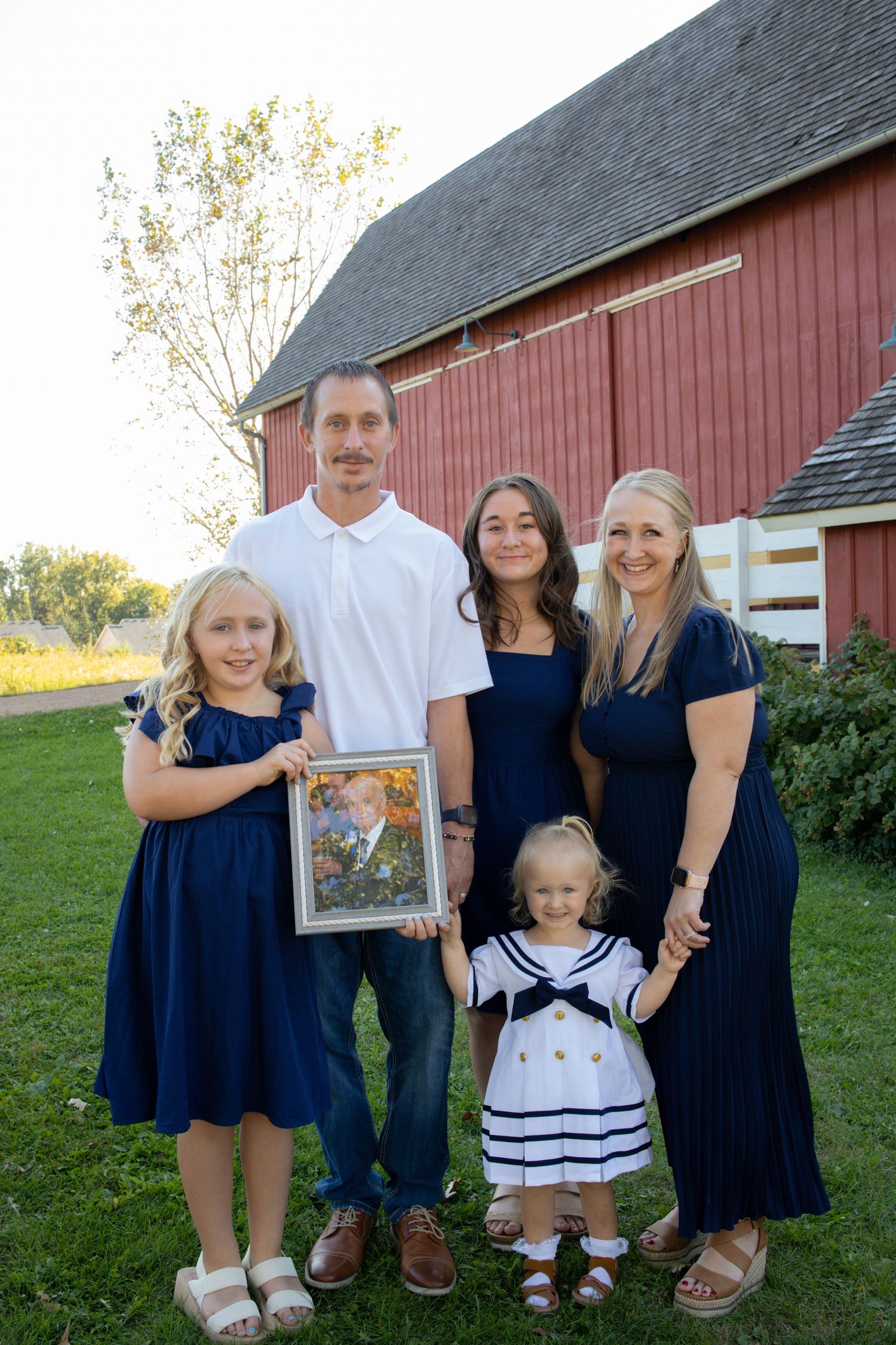 A family wearing coordinated navy blue and white outfits poses together in front of a red barn on a sunny day.