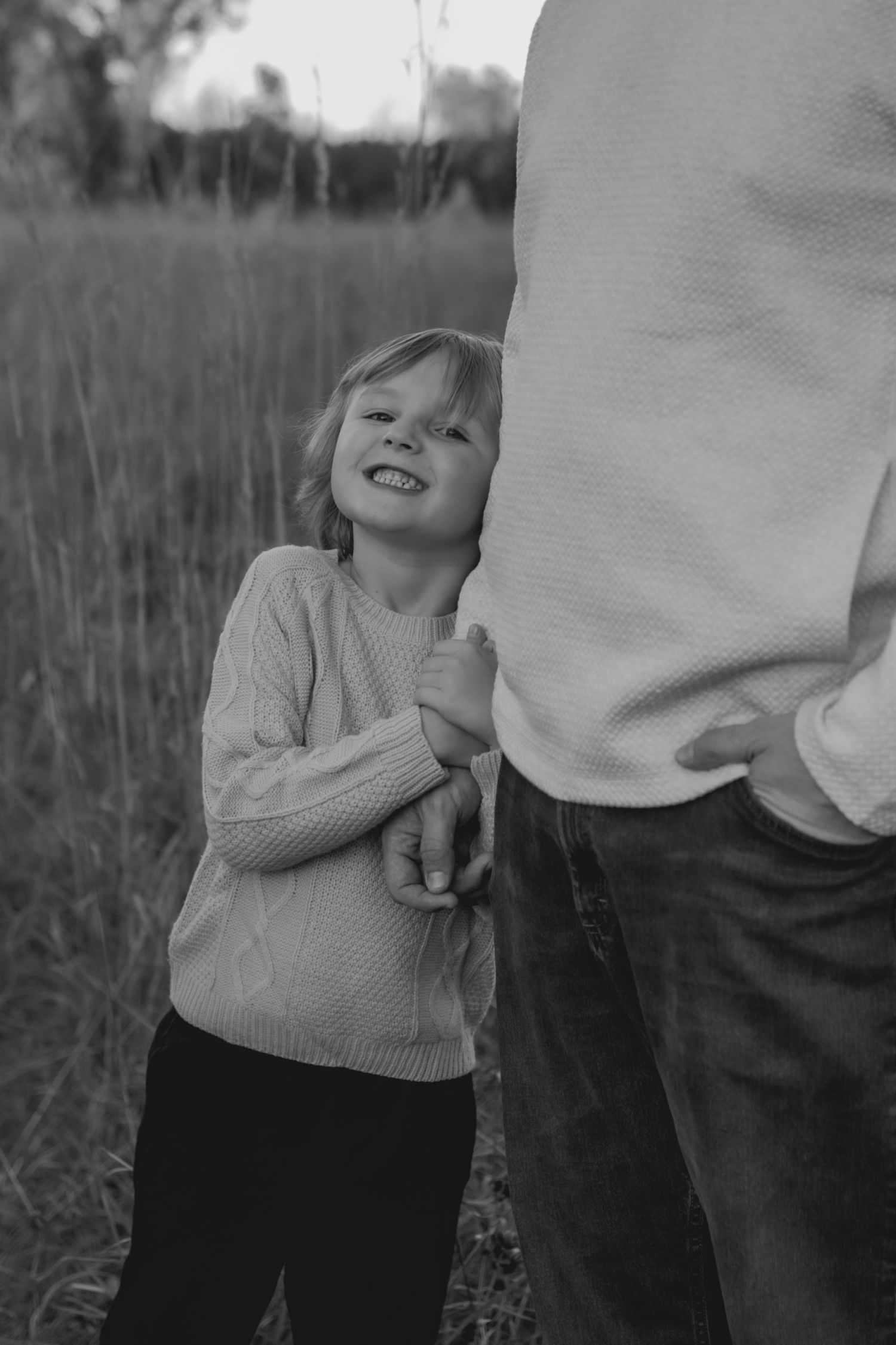 Black and white photo of a child in a sweater holding onto an adult's hand in a grassy field.