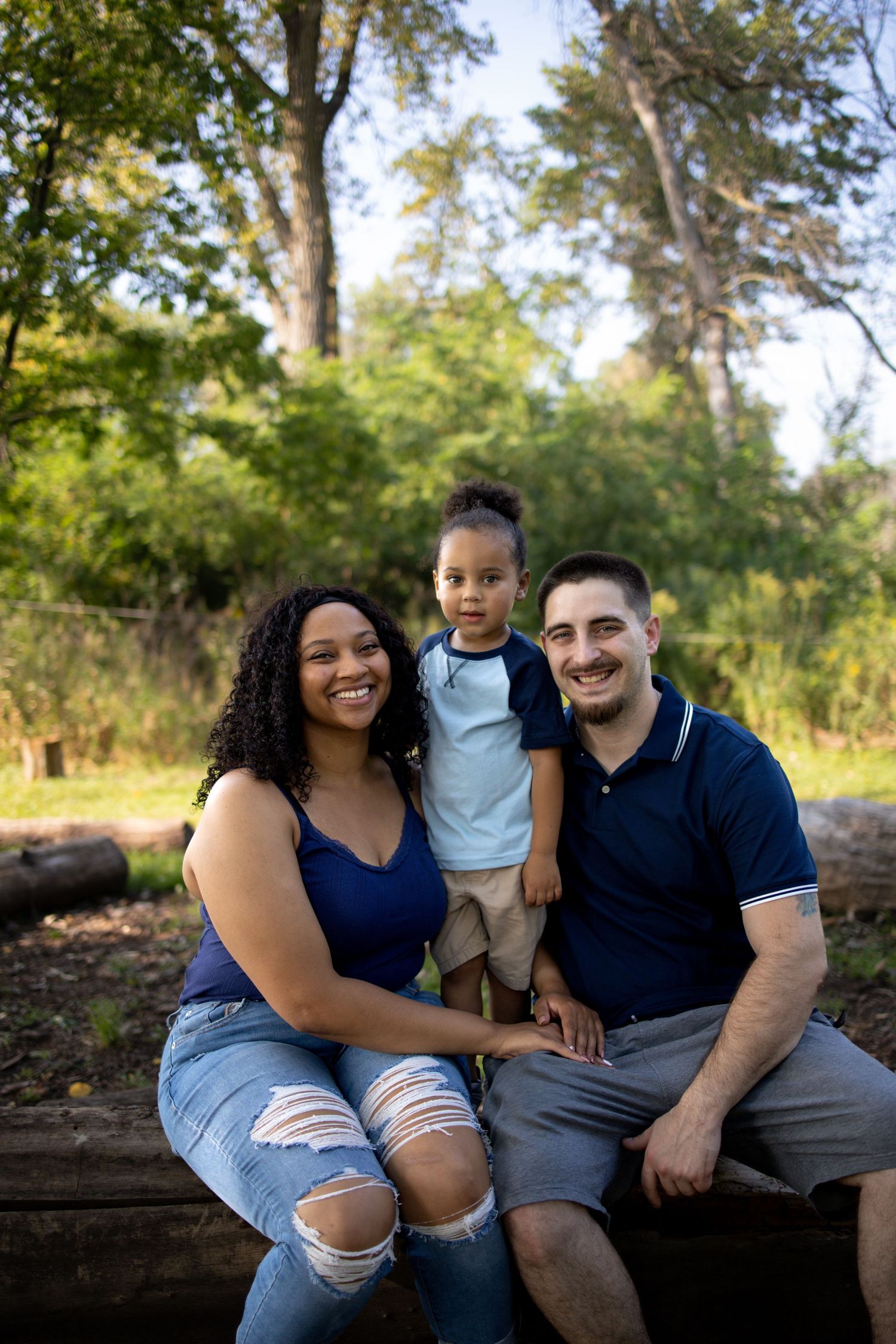 A loving family sits together on a fallen log in a sunlit wooded area wearing coordinated navy blue outfits.