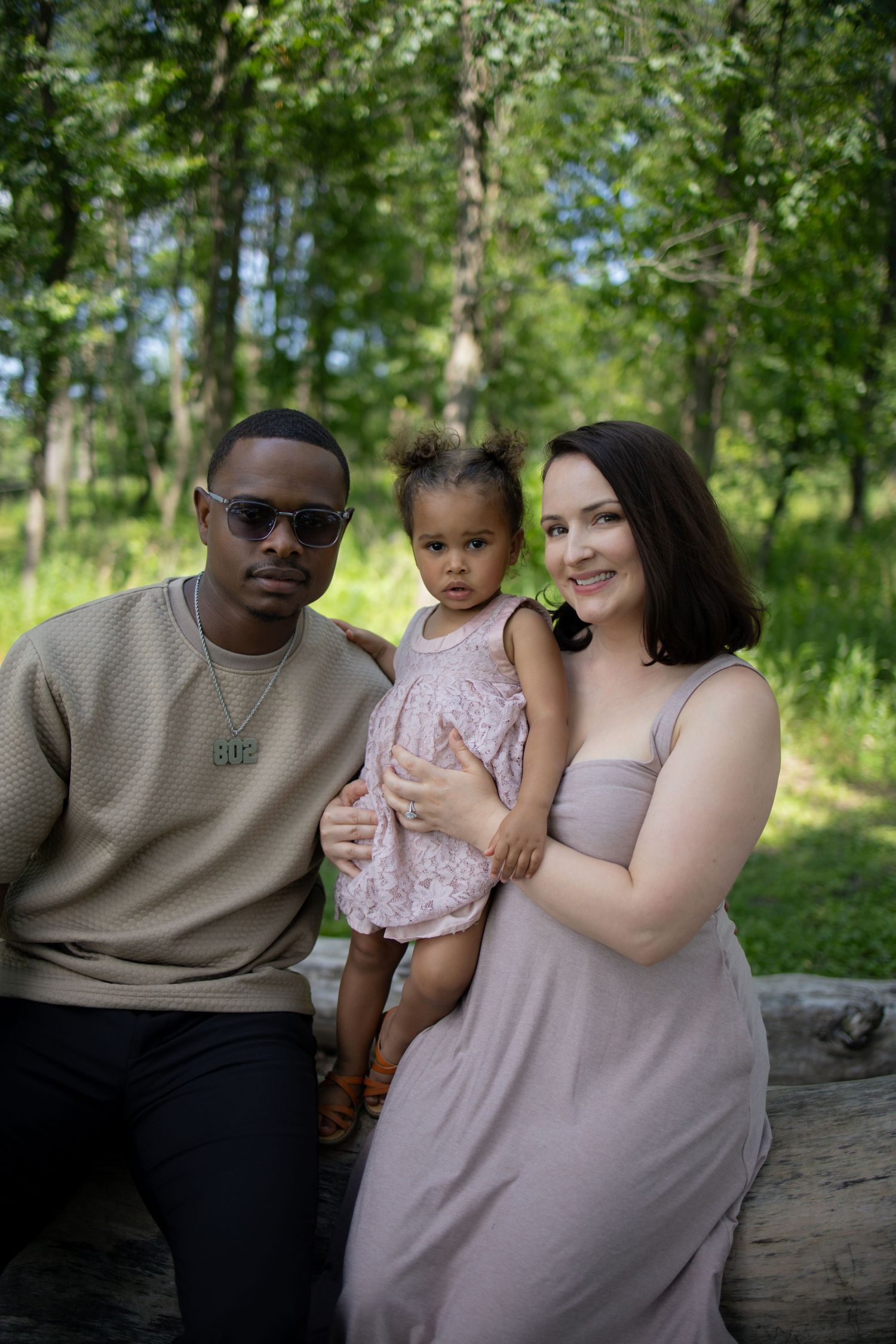 A small family sits together outdoors on a log with a lush green forest background on a sunny summer day.