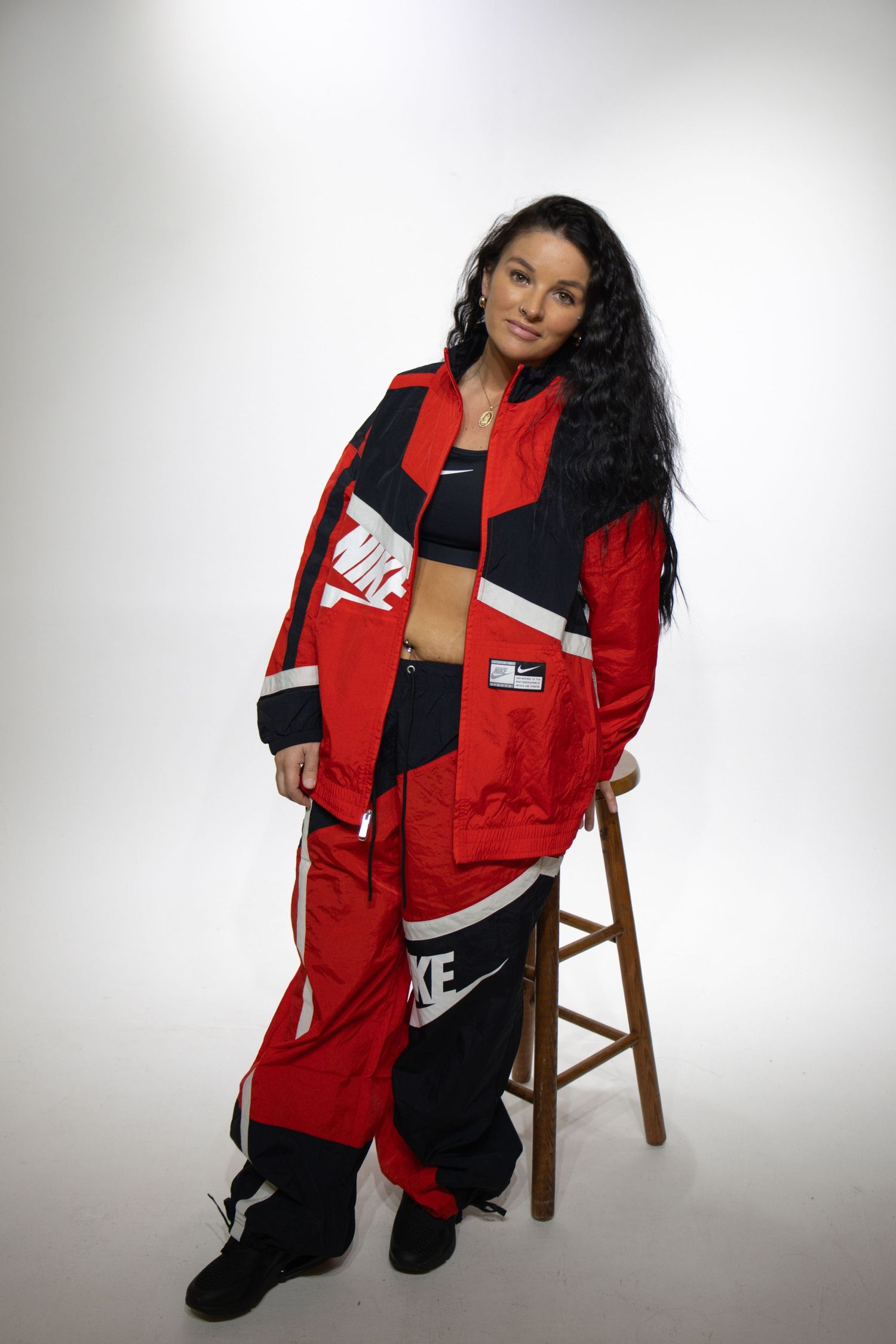 Model in retro red and black Nike sportswear poses on wooden stool against white studio backdrop.