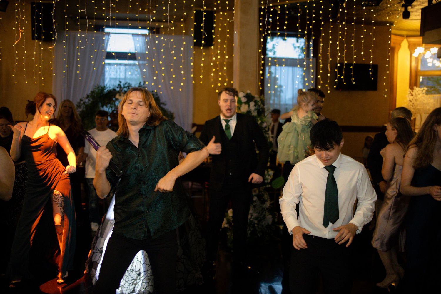Party guests dancing and celebrating at an indoor event with string lights and curtains in the background.