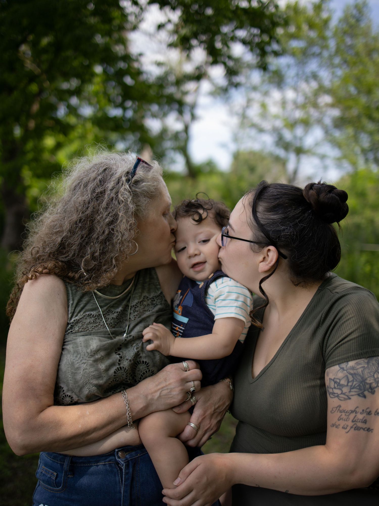 Three generations share a tender family moment outdoors in a natural setting with green trees in the background.