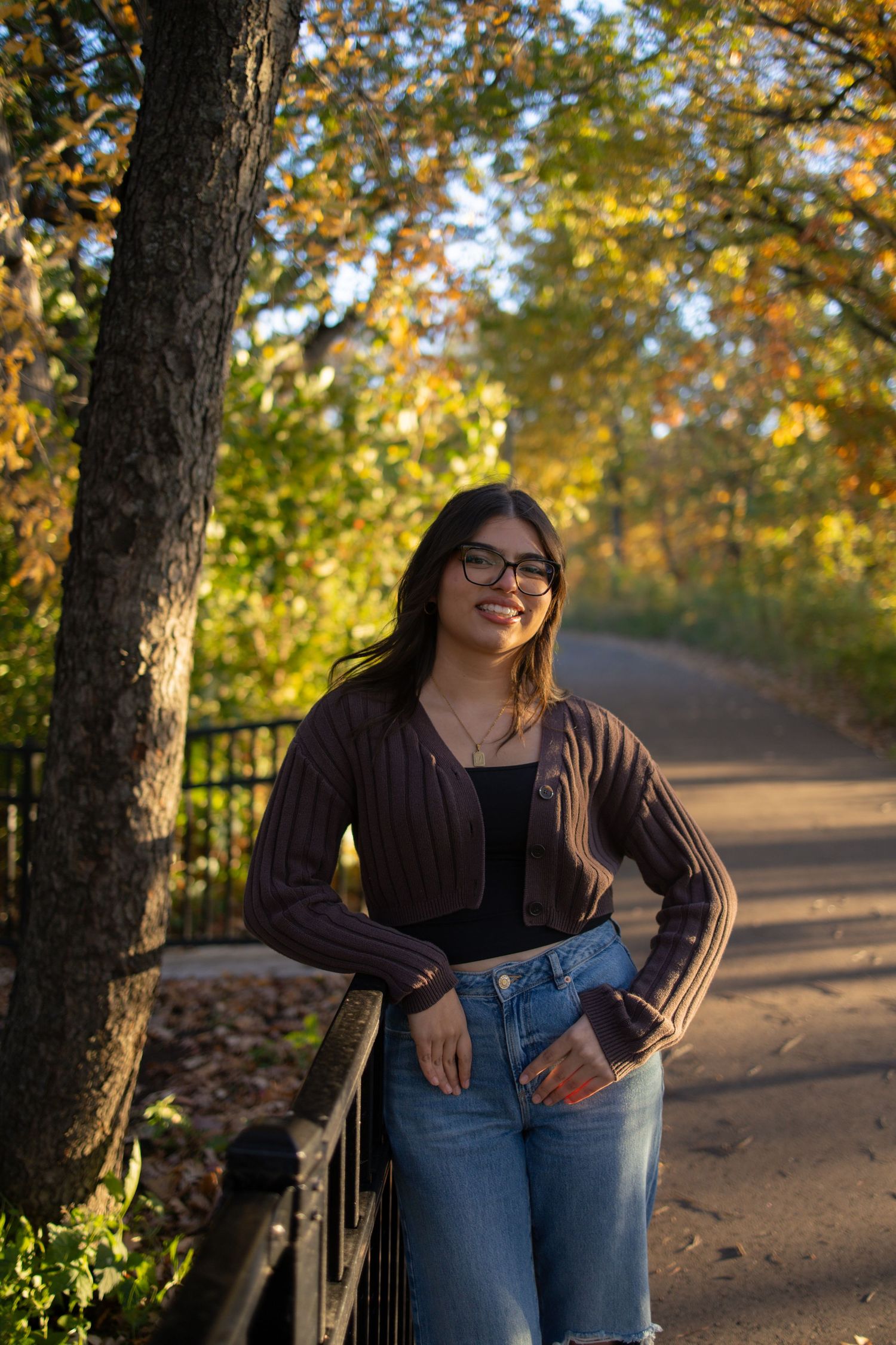 A person in blue jeans and black sweater poses against an autumn backdrop with golden sunlight filtering through trees.