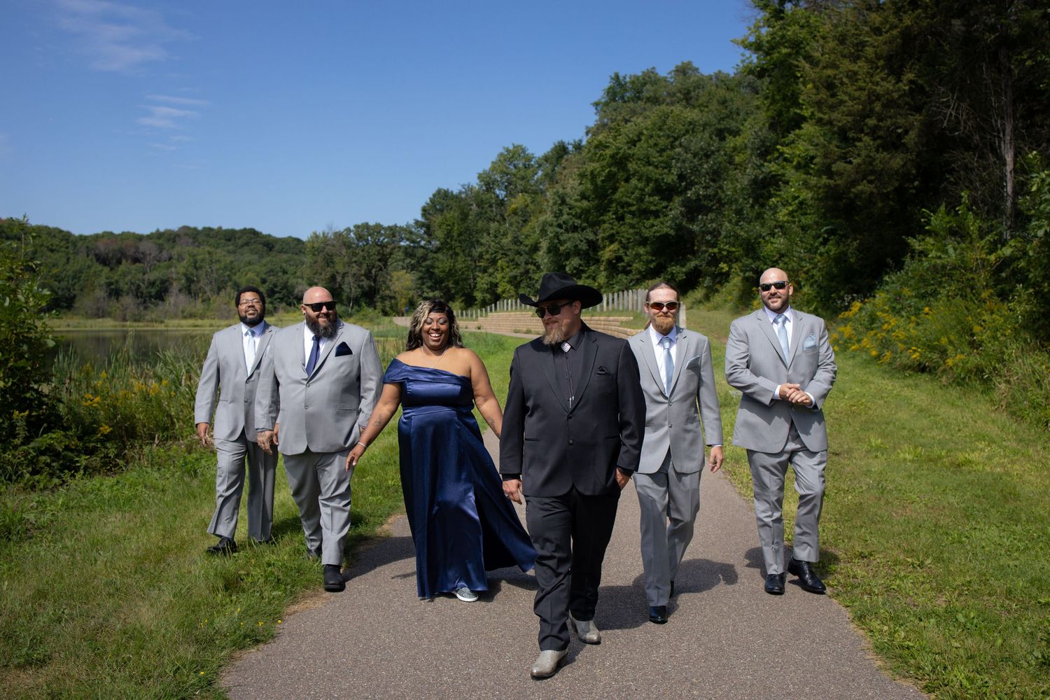 Wedding party members in formal attire walk along a grassy hillside path on a sunny summer day.