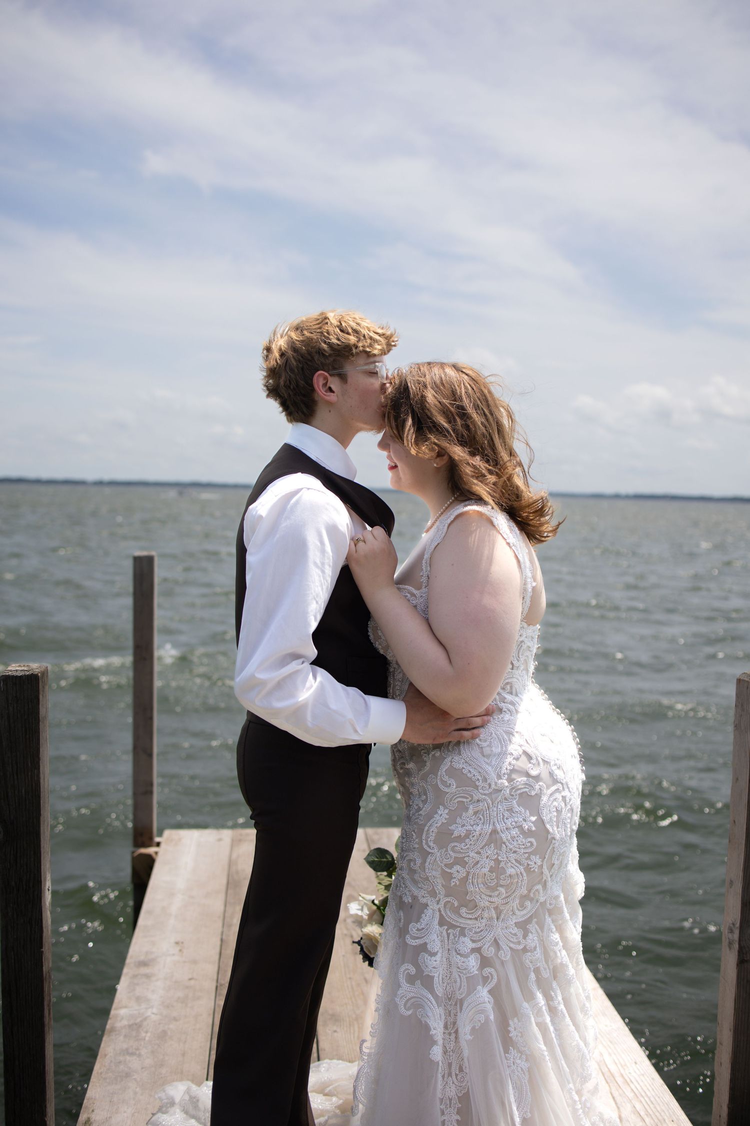 A romantic couple shares a tender moment on a wooden dock over sparkling water under a bright cloudy sky.