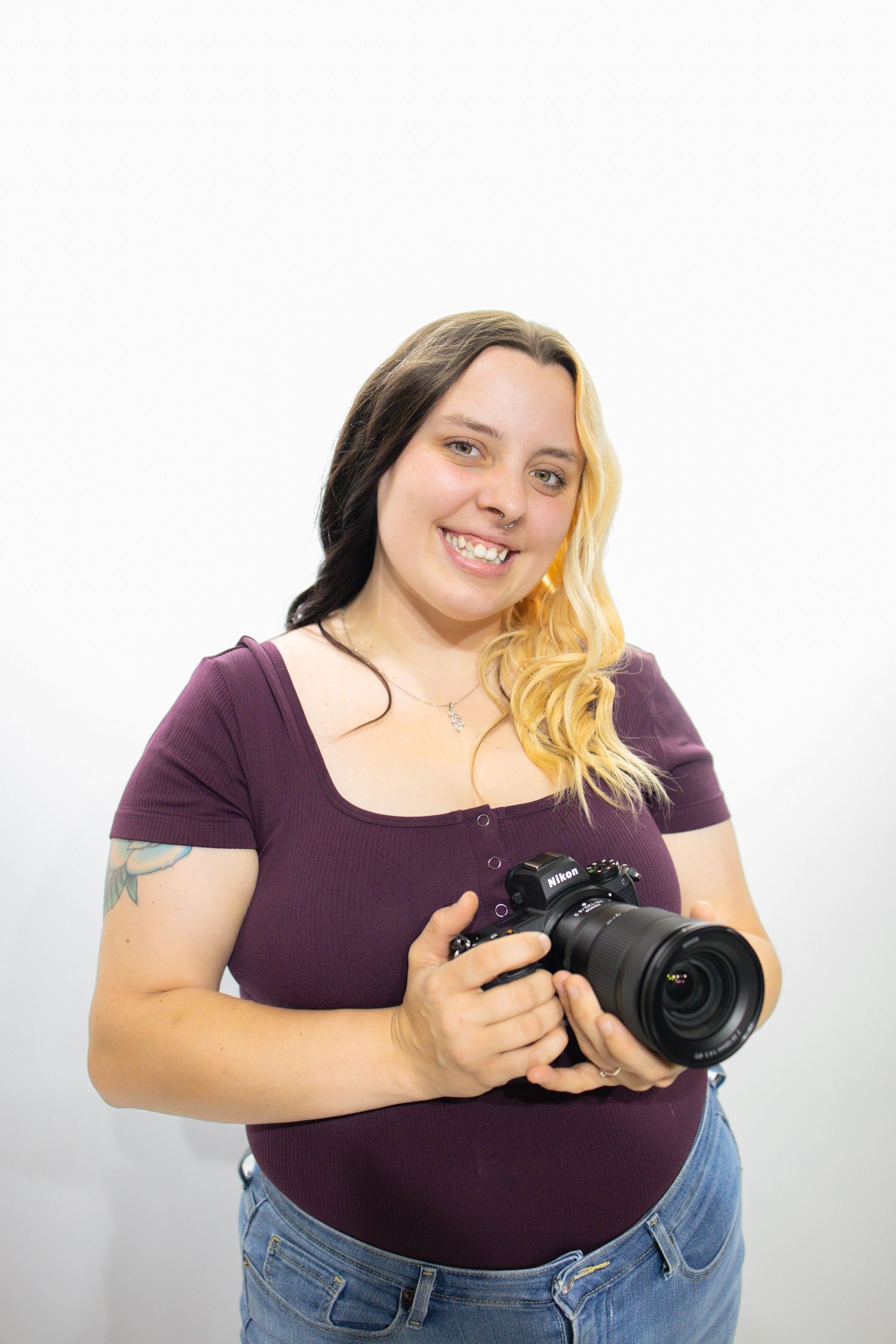 A photographer in a purple shirt holds a professional DSLR camera while standing against a white wall.